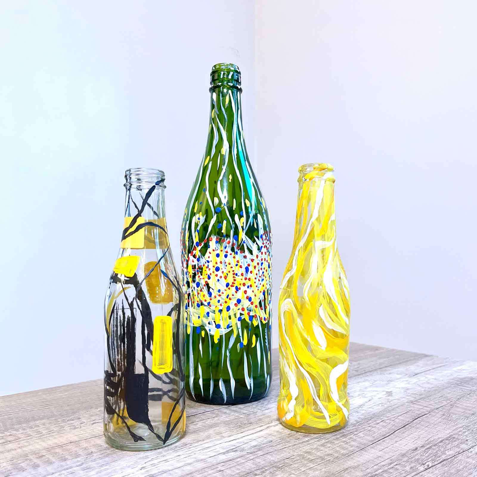 Check out these three vibrant, hand-painted glass bottles sitting on a rustic wooden table. They are perfectly contrasted against a sleek gray backdrop.