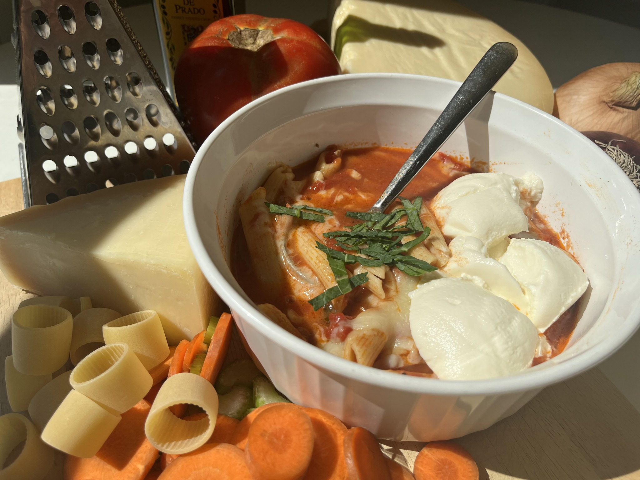 Enjoy a bowl of tomato soup topped with melty mozzarella cheese, beautifully contrasted by vibrant ingredients such as fresh cheese, crunchy carrots, delicious pasta and a traditional grater, all showcased under the glow of natural lighting.