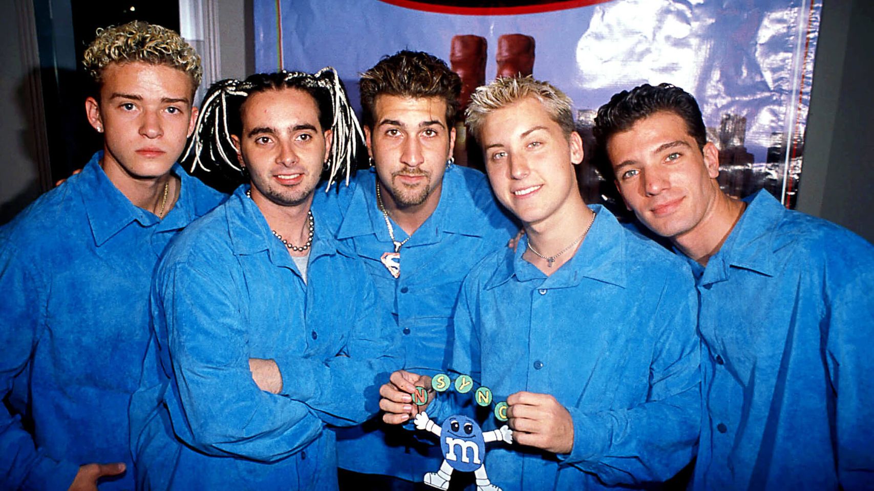 Five cheerful guys in identical blue t-shirts strike a fun pose, with one of them holding a tube of colorful M&Ms.