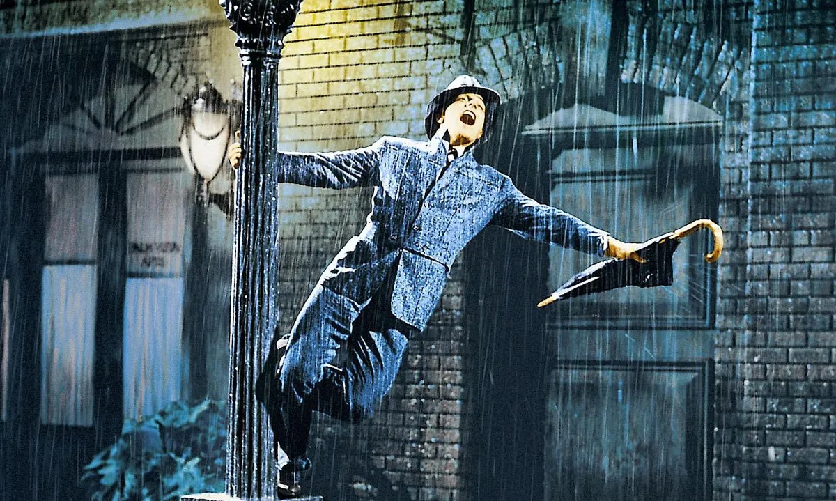 An elegantly dressed man is spotted dancing joyously around a lamp post on a rain-kissed street.