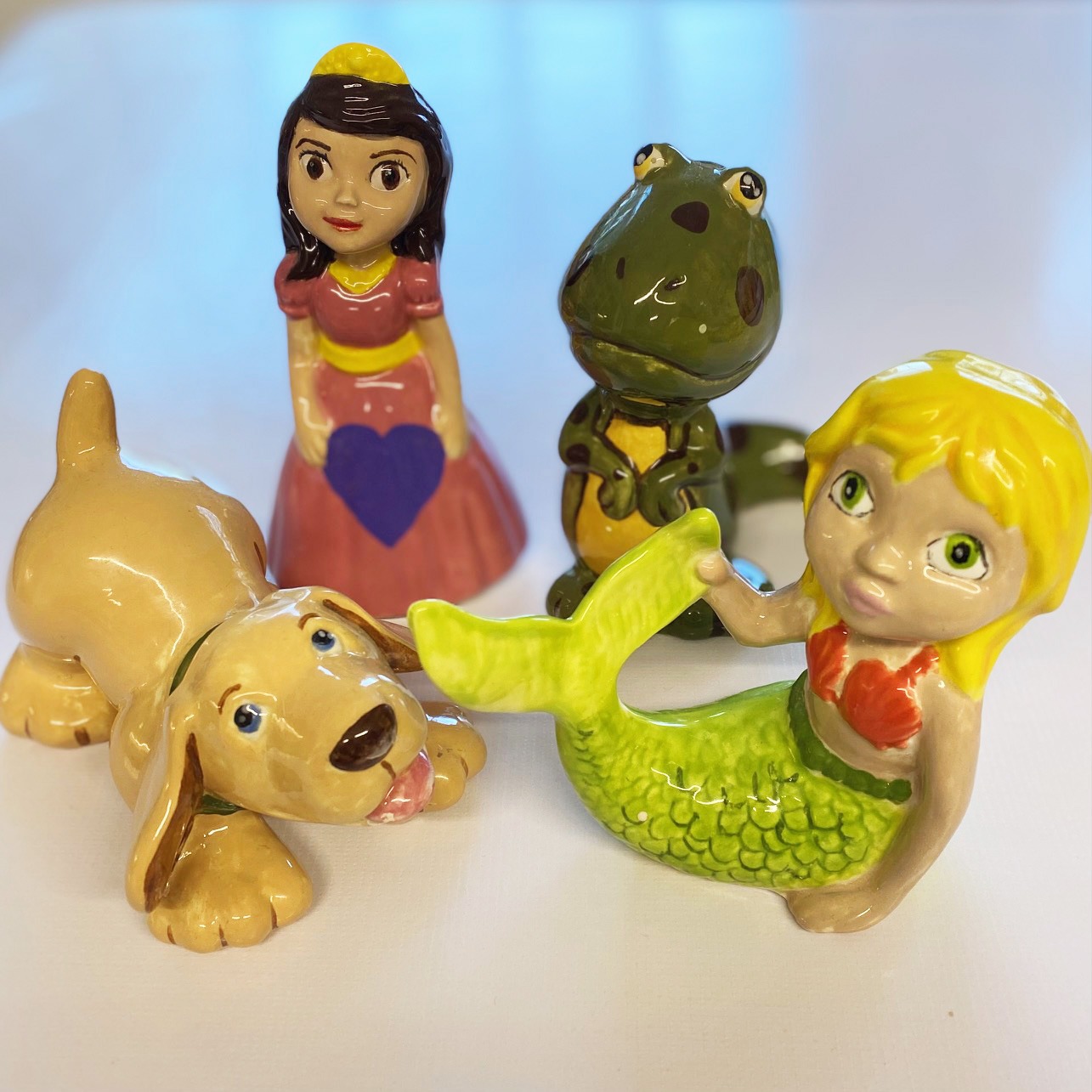 Discover our collection of four unique ceramic figures, each beautifully crafted. Find a charming princess, an adorable frog, a playful dog and an enchanting mermaid all arranged on a clean white background. Perfect for enhancing any living space with personality and character!