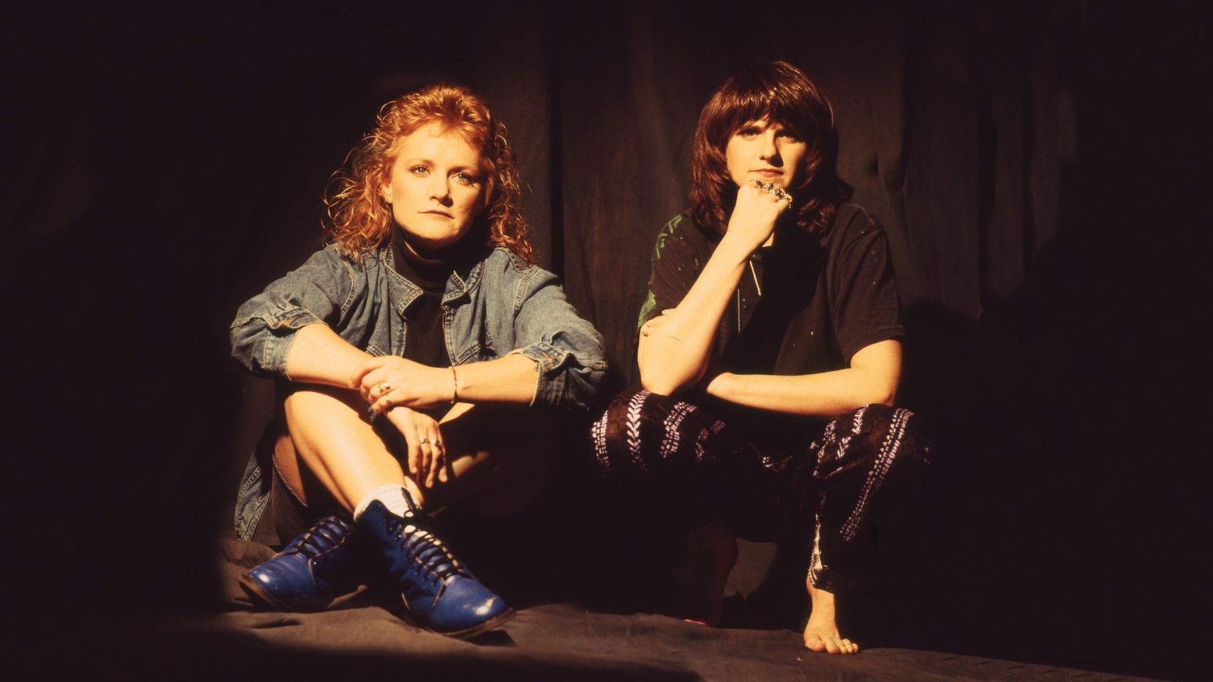 Two women are sitting together in soft lighting. One has curly hair and is dressed casually in a denim jacket and blue shoes. The other woman, sporting straight hair, wears chic black clothing coupled with stylish patterned leggings.