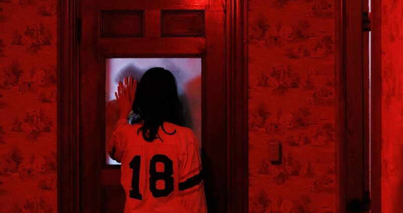 A person in a room bathed in red light is spotted wearing a jersey number 18. They are seen curiously looking through a frosted glass door.