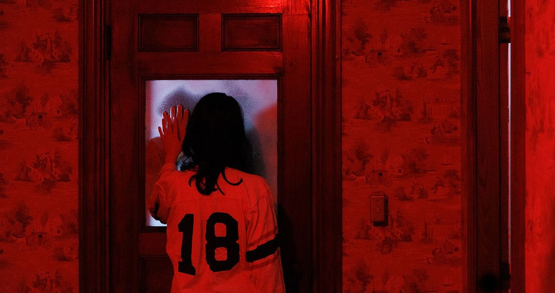 A person in a room bathed in red light is spotted wearing a jersey number 18. They are seen curiously looking through a frosted glass door.