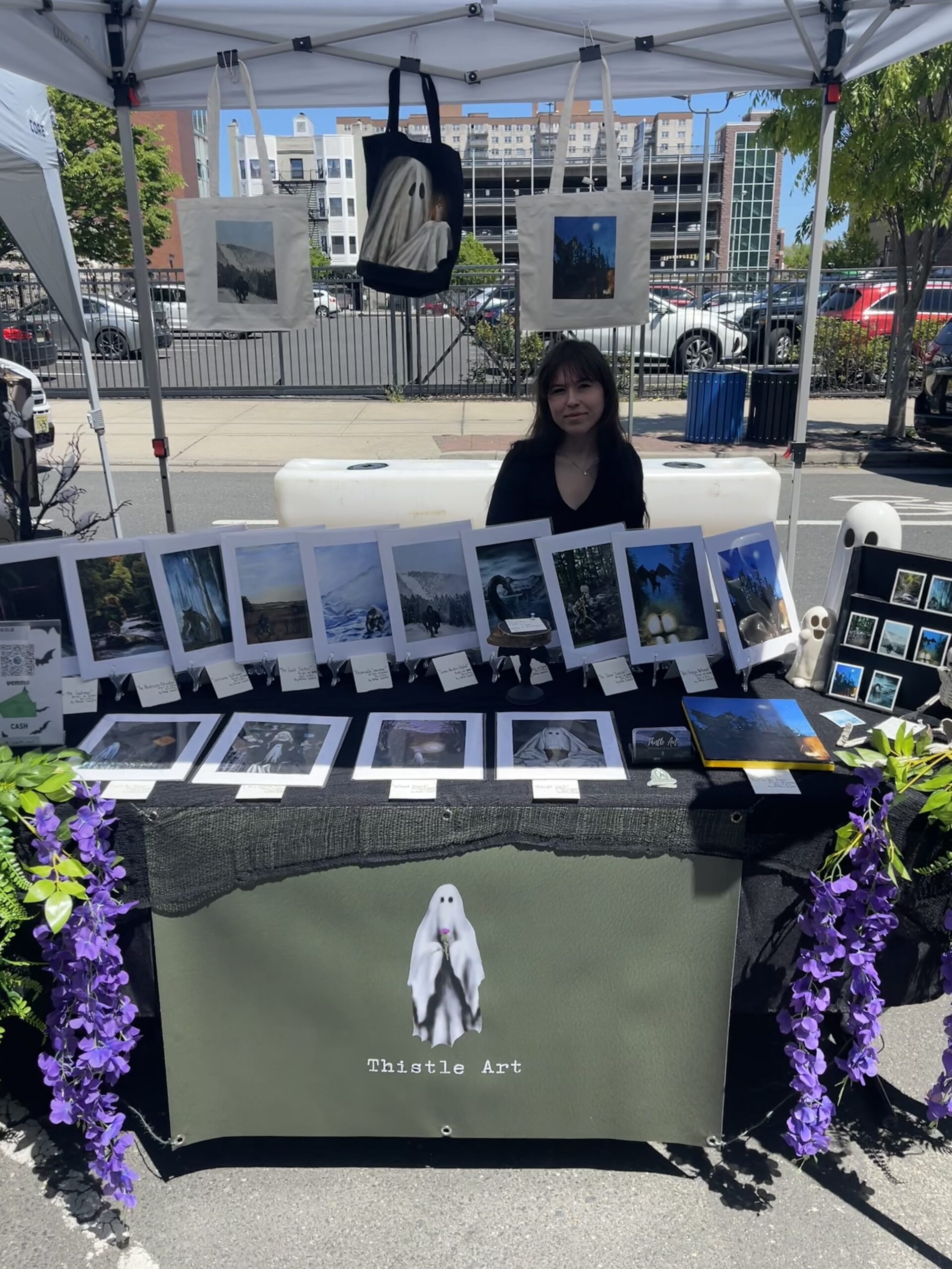 A woman is showcasing photographic art and other creative pieces at an open-air market stall, where you'll also find versatile items like tote bags beautifully displayed overhead.