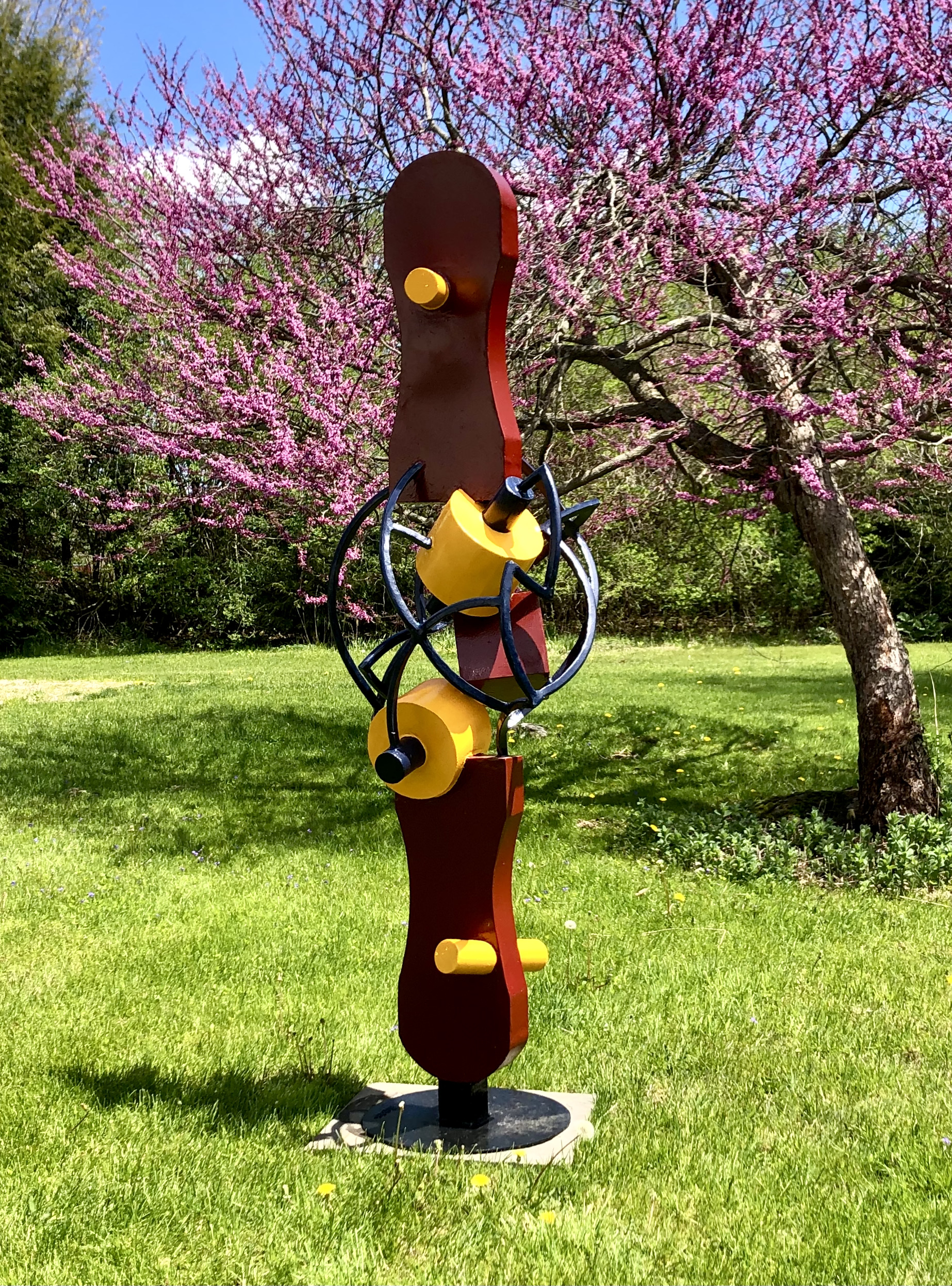 A vibrant, outdoor sculpture with colorful components is artistically presented in a scene of lush green grass and blooming tree.