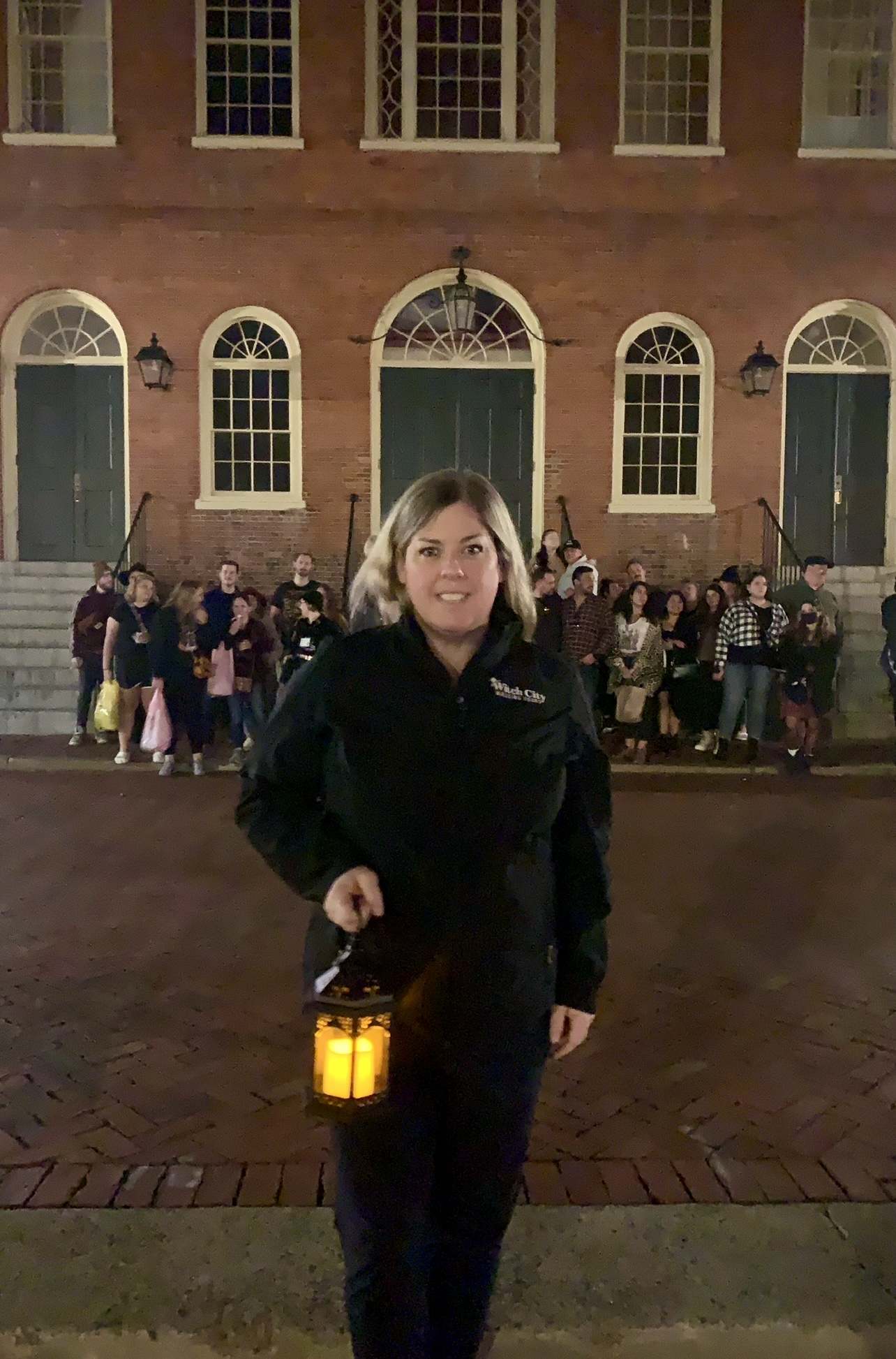 A lady clutching a lantern is positioned before a brick structure during the nighttime, with an assembly of individuals congregated in the shadows behind her.