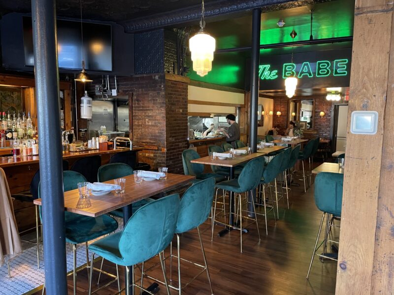 Experience a dining scene with a vibrant flair at our restaurant. Revel in the appealing blend of teal cushioned chairs and earthy wooden tables while appreciating the trendy bar area. Be captivated by the luminous neon signage that boasts our unique title - "the babe.