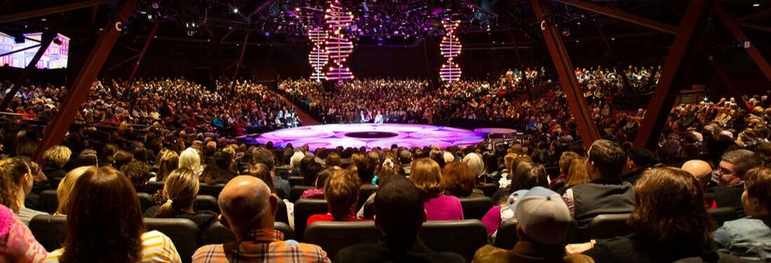 Audience positioned in a theater featuring a round stage and brightly lit designs.