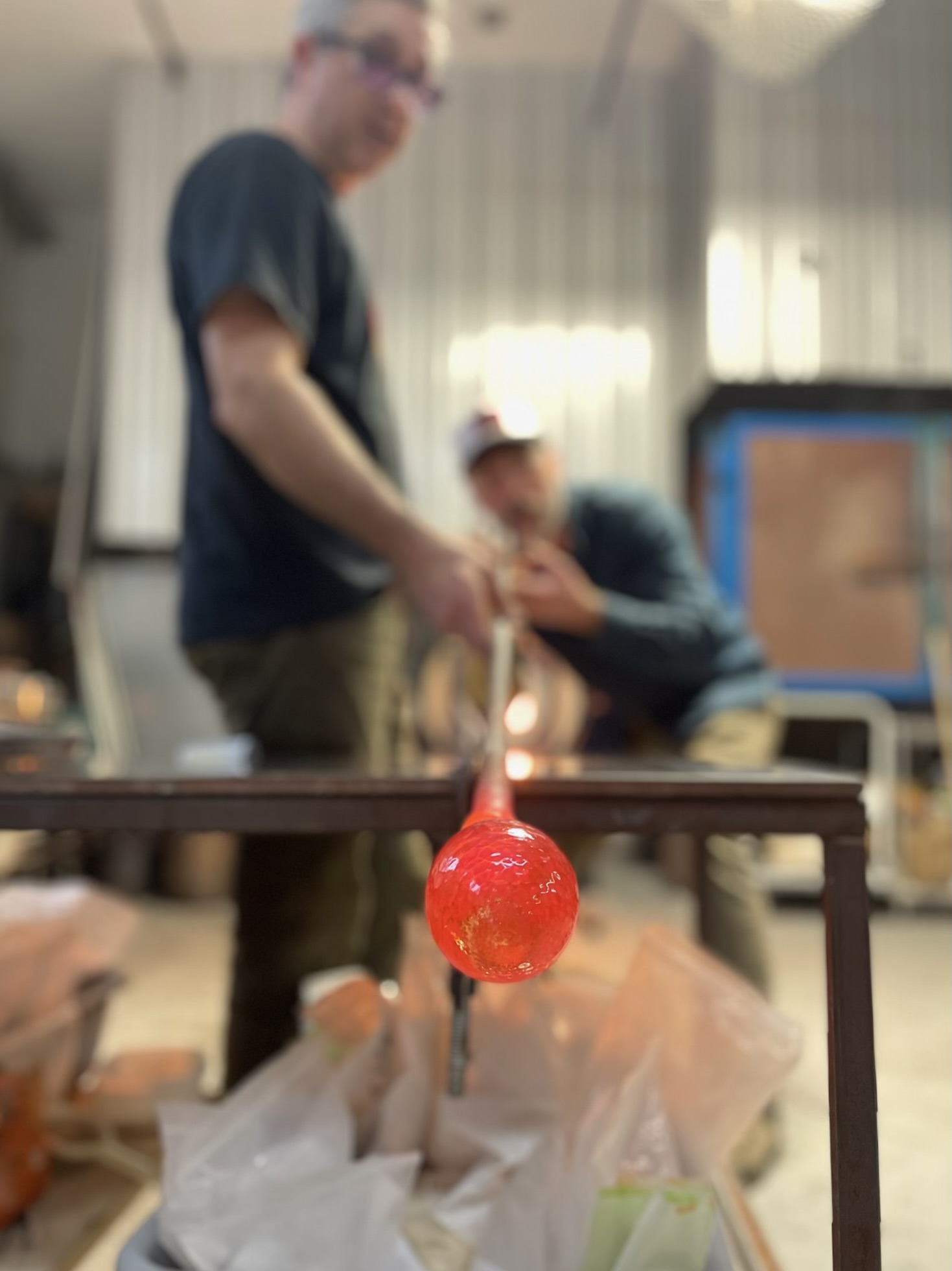 A professional glassblower skillfully crafting a fiery molten glass piece on a rod, while an individual watches from the soft focus backdrop of a bustling workshop.