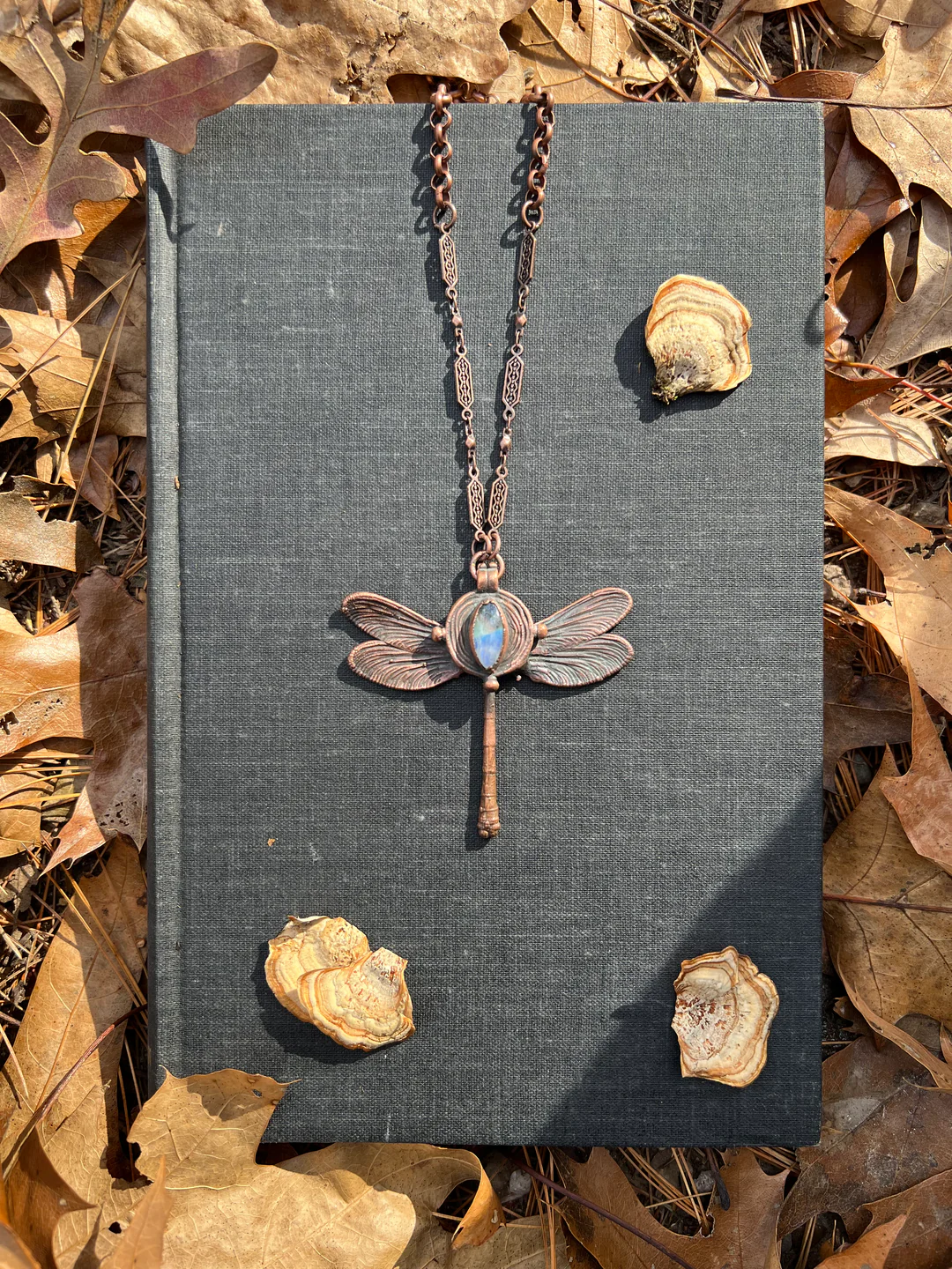 Discover the glow of our dragonfly pendant necklace, beautifully showcased against a charming gray book, amidst the rustic allure of autumn leaves and pieces of tree bark.