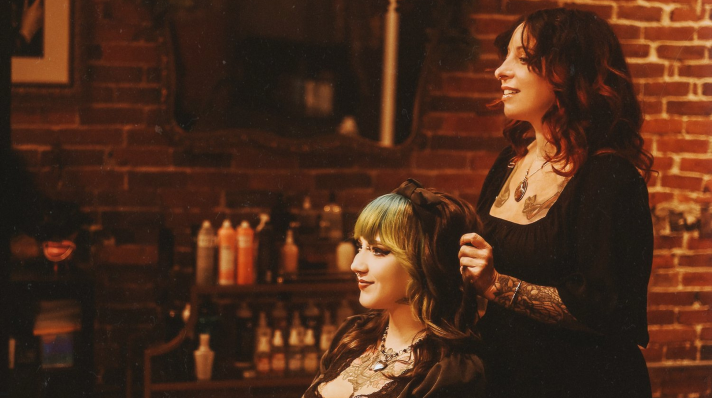 A tattooed hairstylist expertly crafts a client's look in a cozy salon with charming brick walls and an array of hair products neatly displayed in the background.