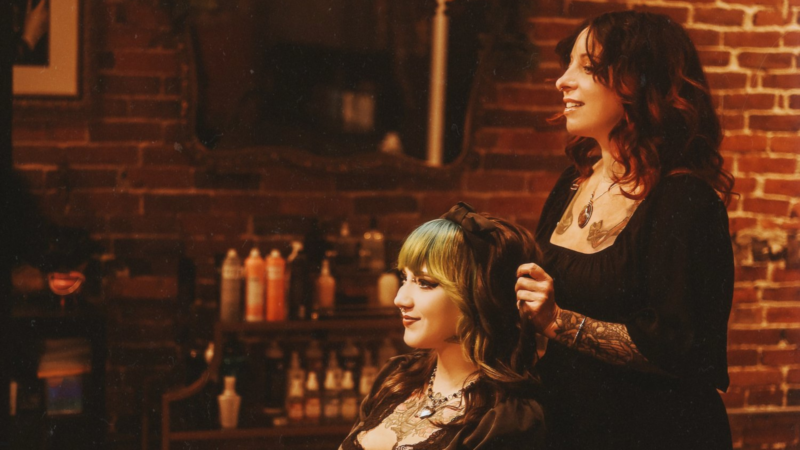 A tattooed hairstylist expertly crafts a client's look in a cozy salon with charming brick walls and an array of hair products neatly displayed in the background.