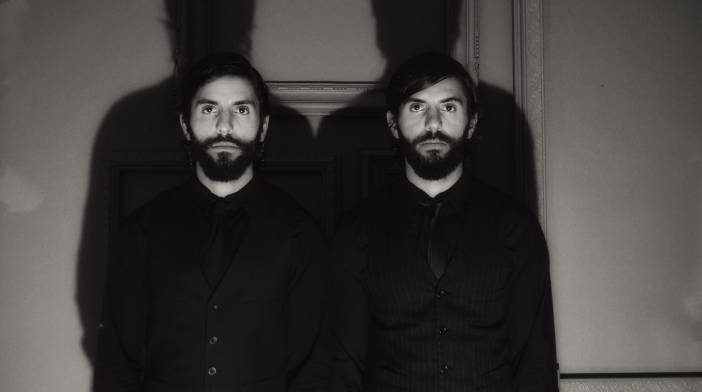 In a moody, dimly lit room at The Black Veil Studio, two bearded men stand shoulder to shoulder. Their dark attire perfectly complements the mysterious ambiance, with their shadows creating an intriguing silhouette on the wall behind them.