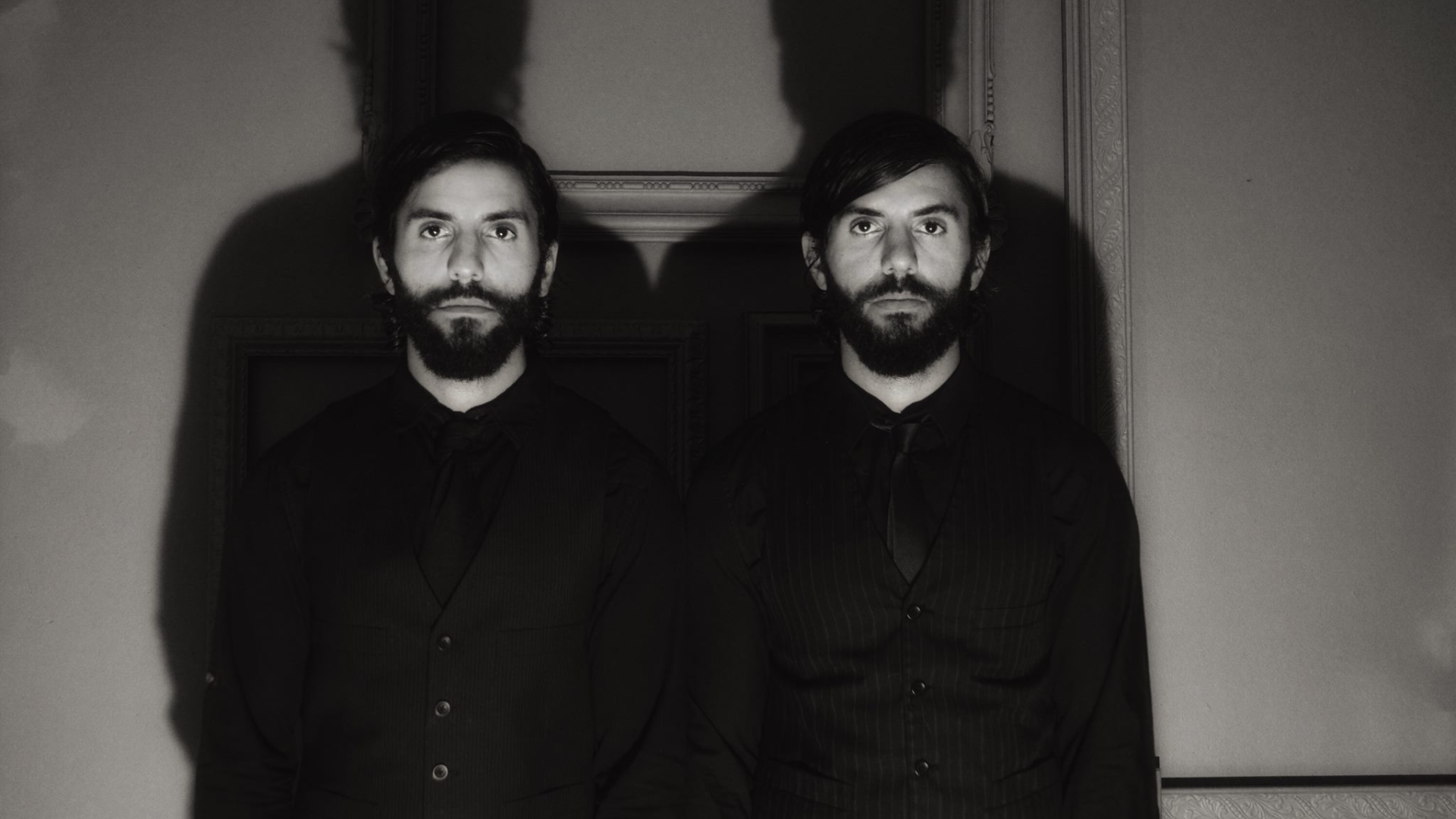 In a moody, dimly lit room at The Black Veil Studio, two bearded men stand shoulder to shoulder. Their dark attire perfectly complements the mysterious ambiance, with their shadows creating an intriguing silhouette on the wall behind them.