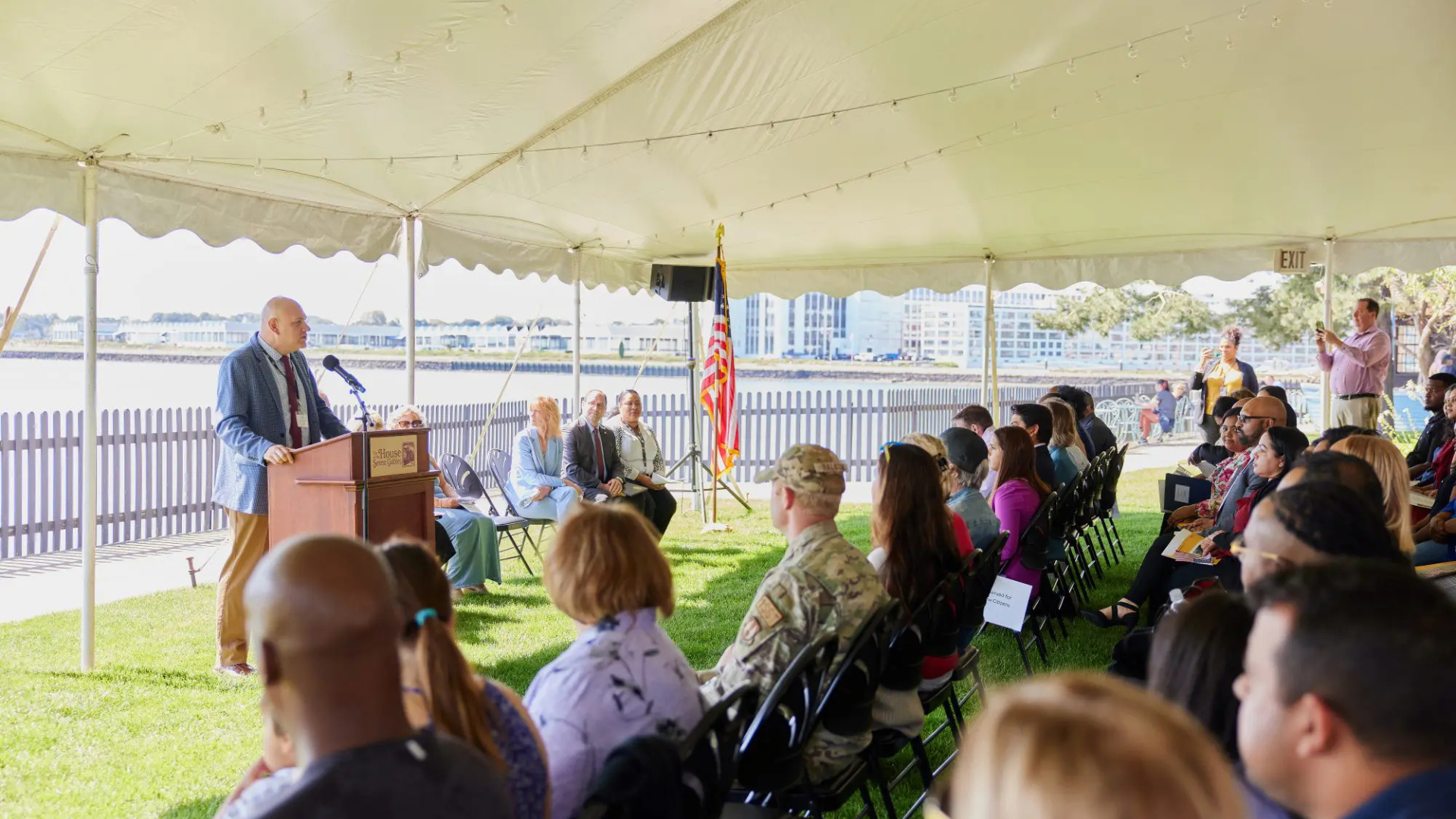 A speaker addresses an engaged audience under a spacious white tent set against a scenic waterfront backdrop. Among the attendees are military personnel, highlighting the event's significance.