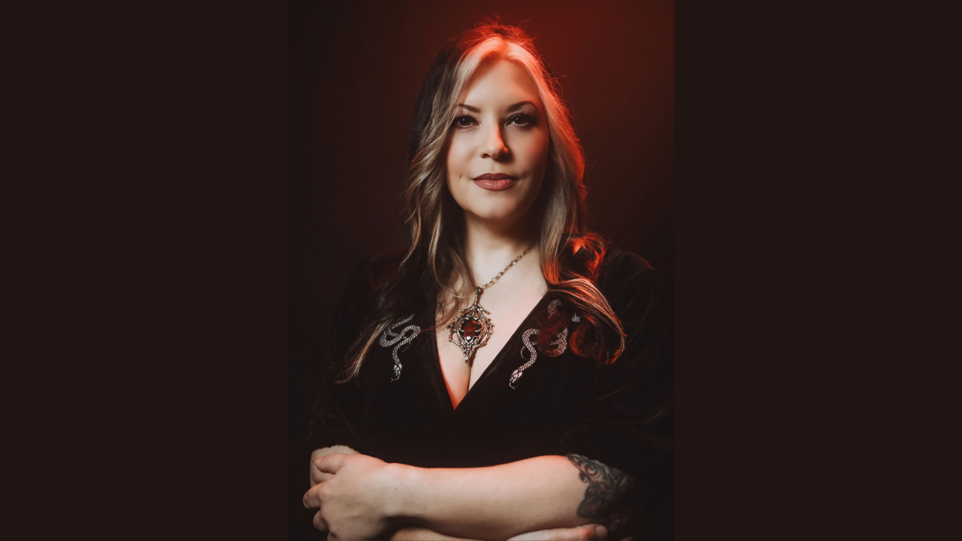 Spotlight on a Member: A woman showcases her long, flowing hair styled with Moon Baby Hair Salon's signature touch. Her forearm tattoo adds personality to her look, as she confidently stands with crossed arms. Sporting a pendant necklace and dark top, she's set against an intriguing red glow backdrop that draws you in.