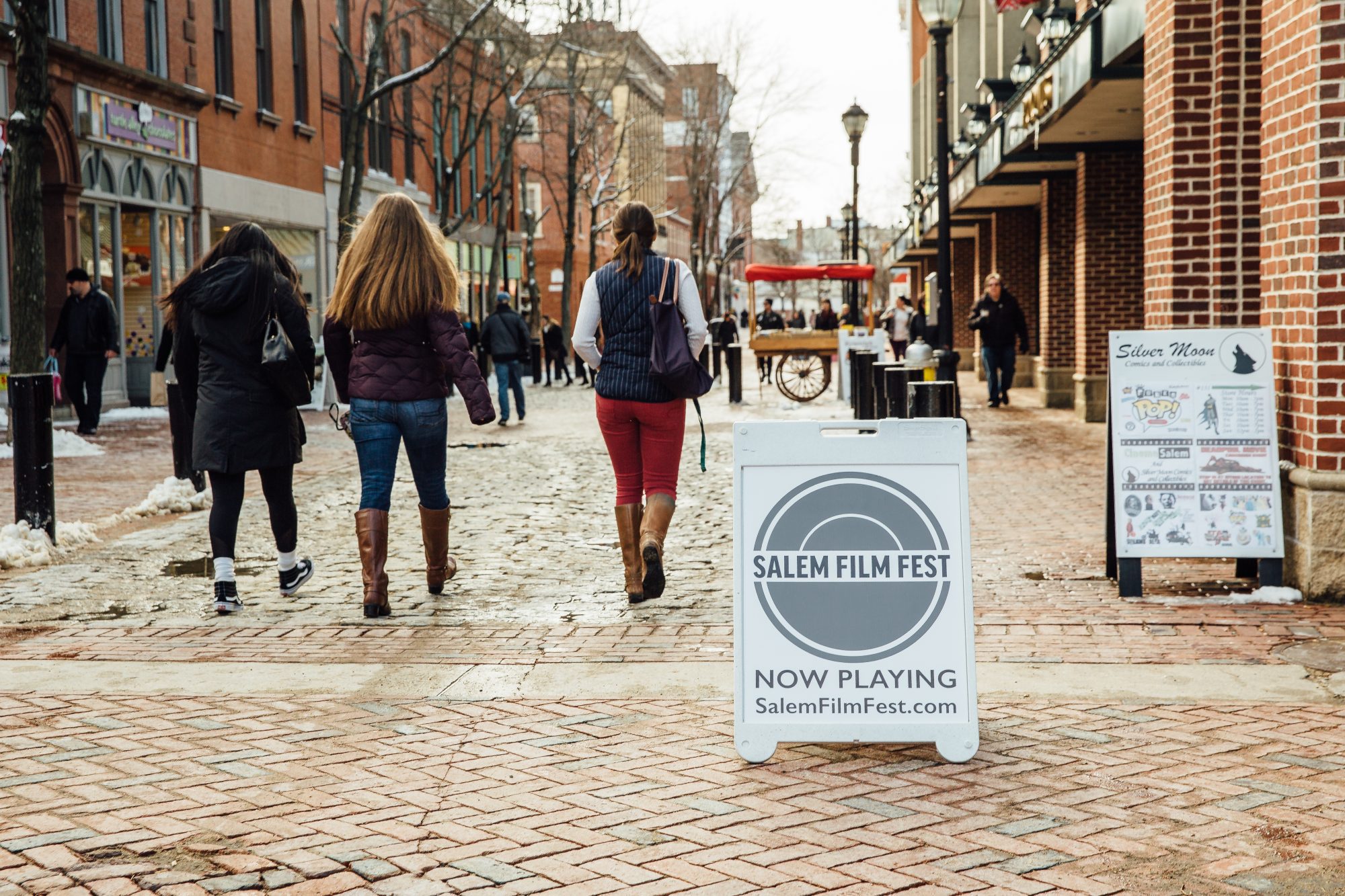 Pedestrians stroll down a brick path, passing a sign heralding the Salem Film Fest.