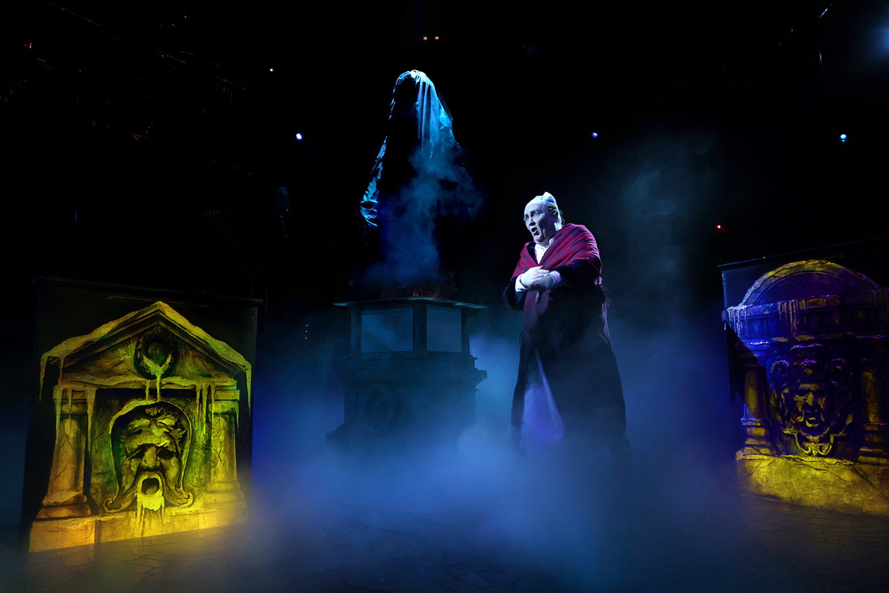 A person dressed in red strikes a dramatic pose on stage, enveloped by fog and glowing statues, while a cloaked figure looms in the background. This captivating scene captures attention and draws viewers into its mystery.