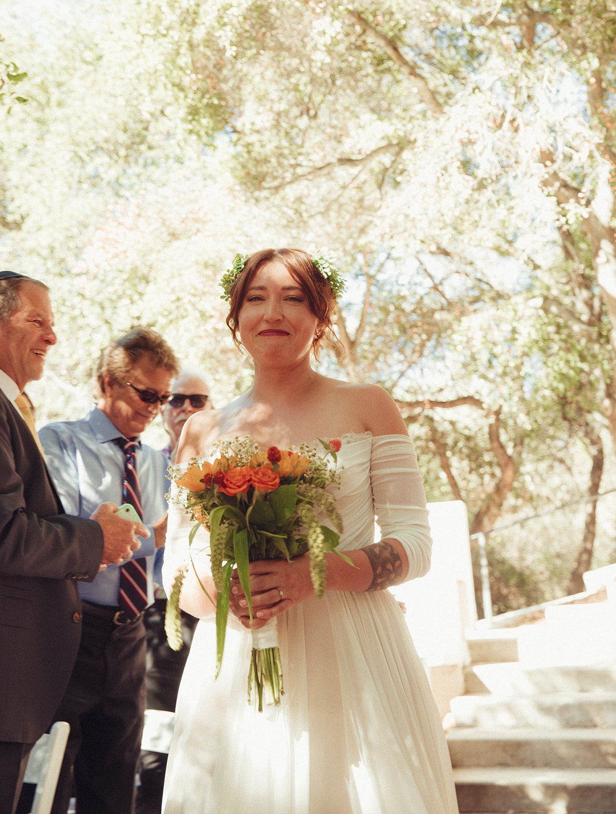 Capture the perfect outdoor wedding moment: a radiant bride in a stunning white dress with delicate floral patterns, joyfully holding her bouquet. Set against a backdrop of lush trees and surrounded by loved ones, this scene is full of natural beauty and happiness.