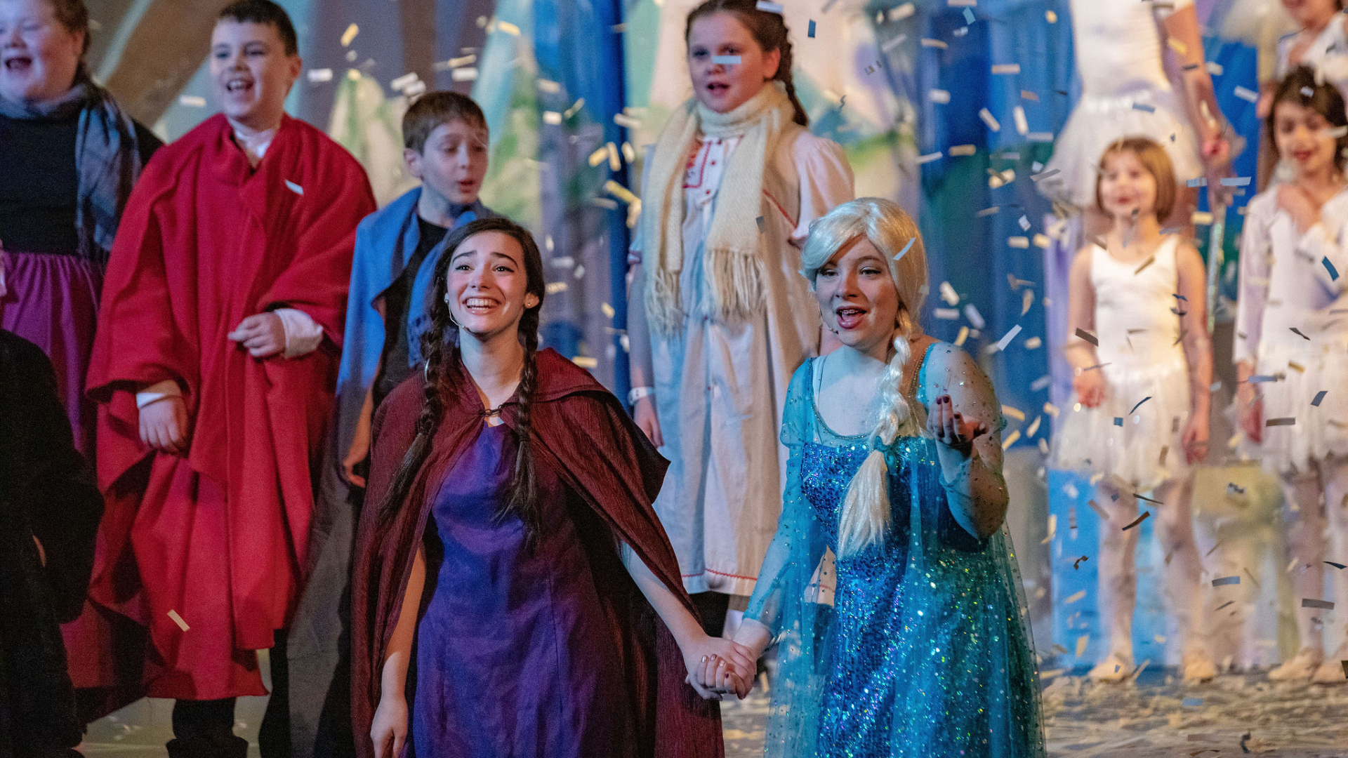 Immerse yourself in the vibrant spectacle of performers in costume on stage, as confetti rains down. Picture a character in a blue dress with a braided wig holding hands with another clad in a burgundy cloak, all amid an ensemble dressed in a lively array of outfits. This scene vividly captures the theatrical flair and community spirit, much like "Meet the Member: Salem YMCA.