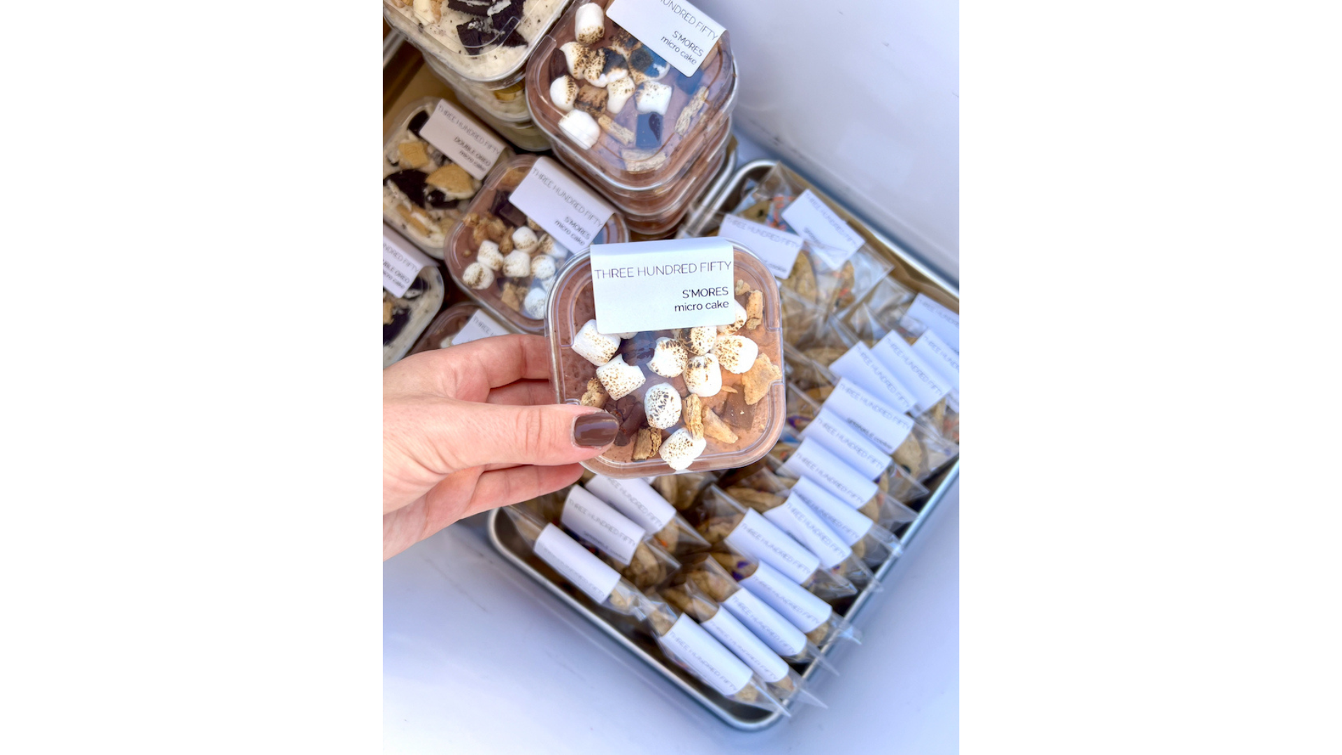 A hand reaches for a container marked "Three Hundred Fifty," highlighting delicious mini cakes surrounded by an enticing assortment of packaged pastries.