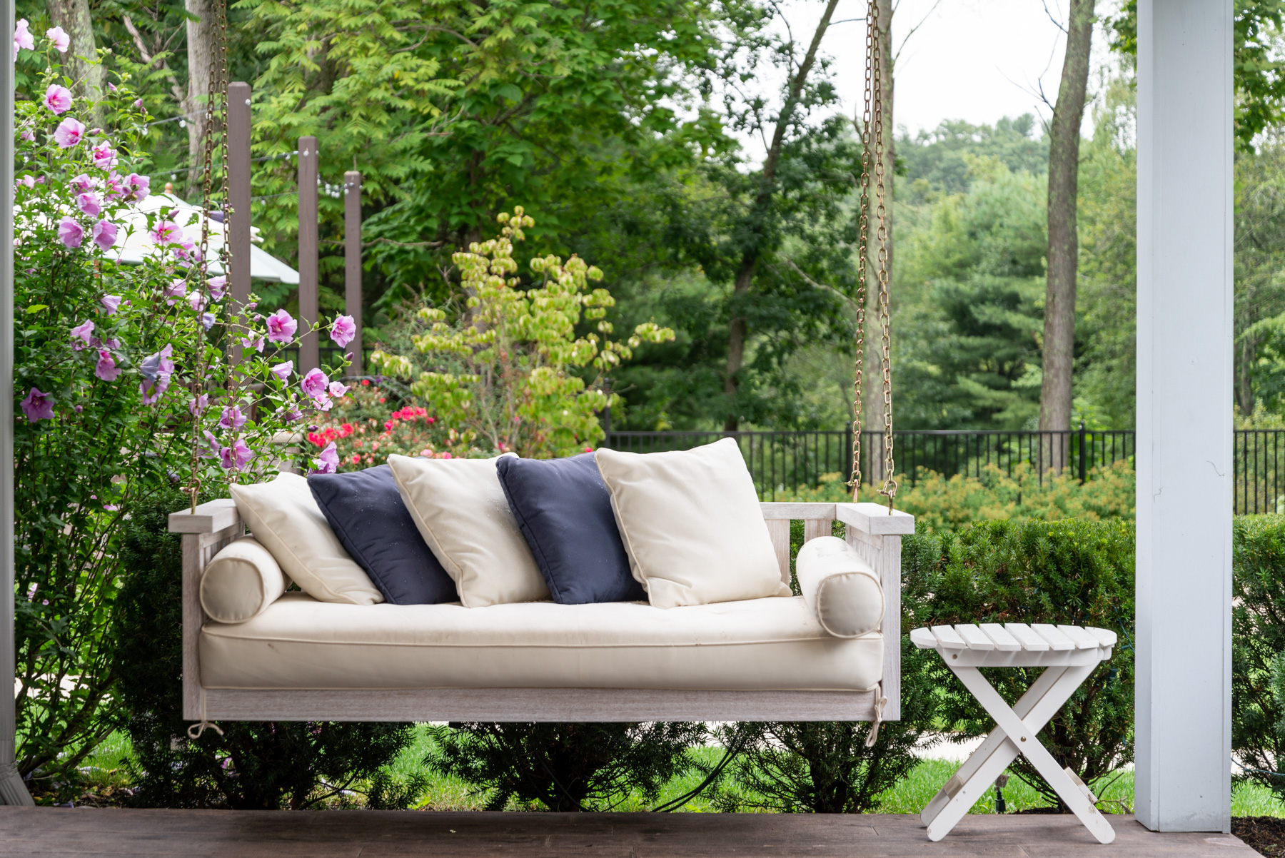 Transform your outdoor space into a cozy retreat with our elegant white porch swing. Complete with plush cushions, it's the perfect spot to unwind. Paired with a convenient white folding table and nestled among vibrant greenery and blooming shrubs, this setup offers a serene escape right at home.