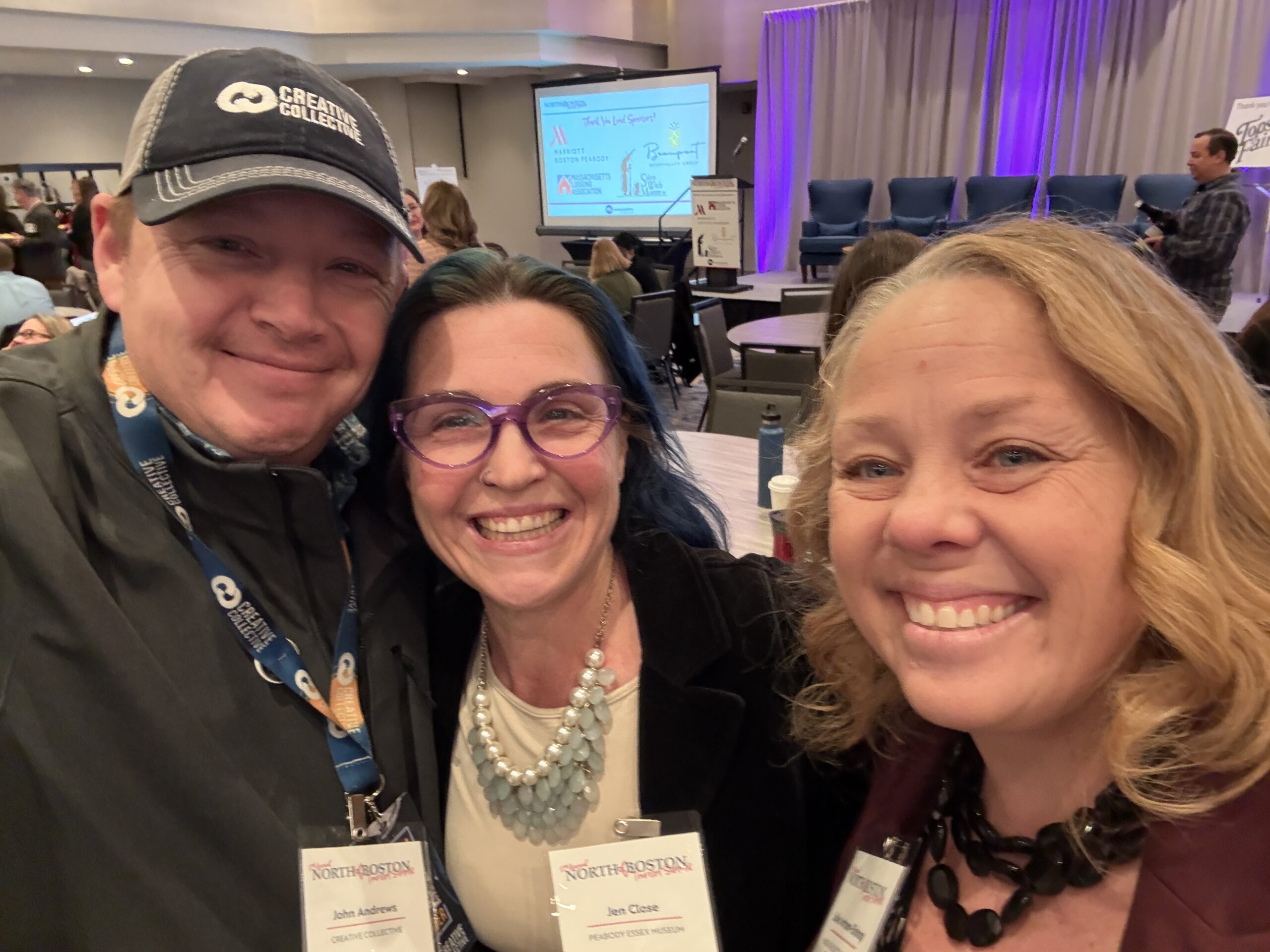 Three people share smiles, capturing a selfie at an indoor event. Behind them, you can see an audience seated and ready for the presentation on stage.