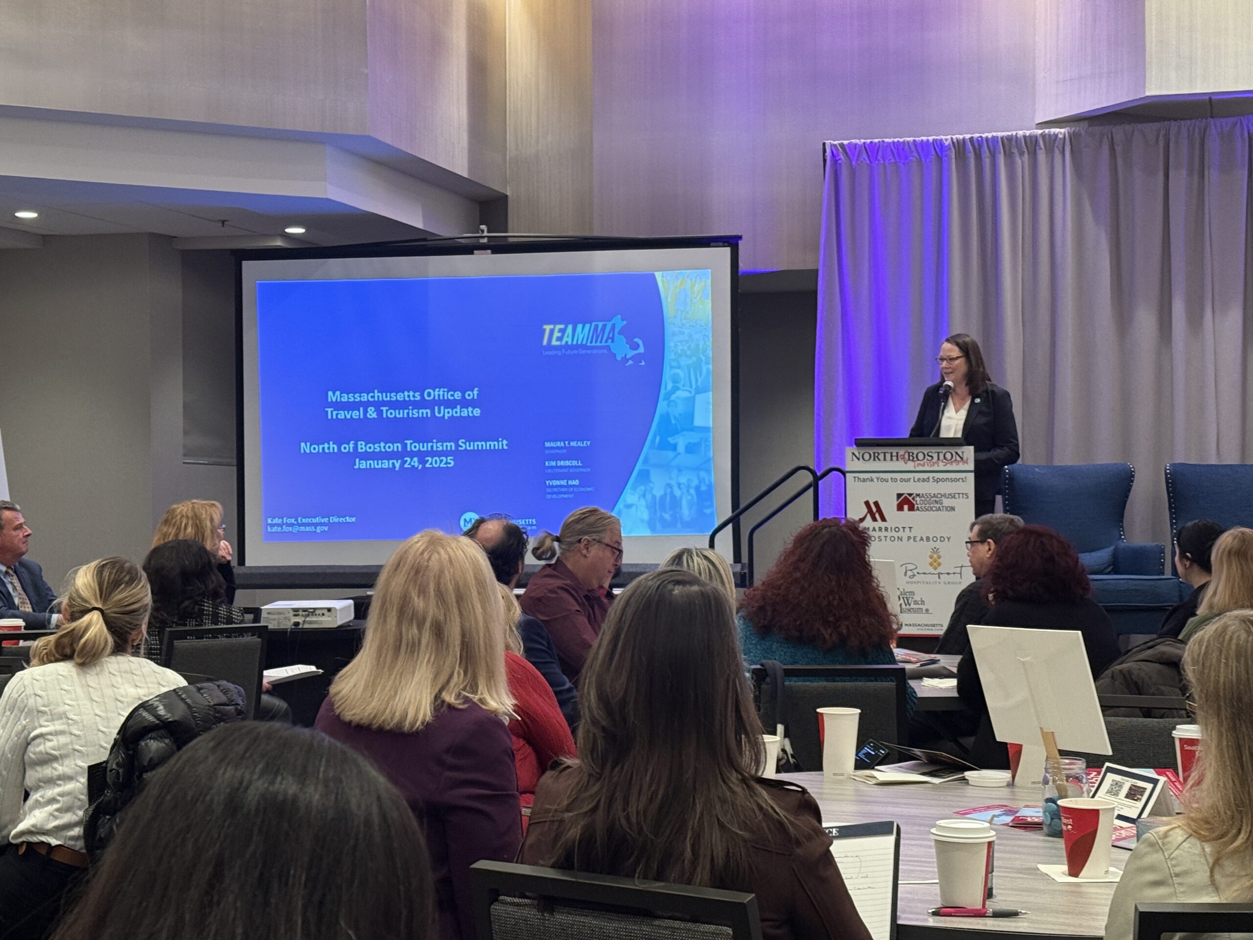 A woman takes center stage at the North of Boston Tourism Summit, captivating attendees seated around tables. A screen prominently showcases the event name and date.
