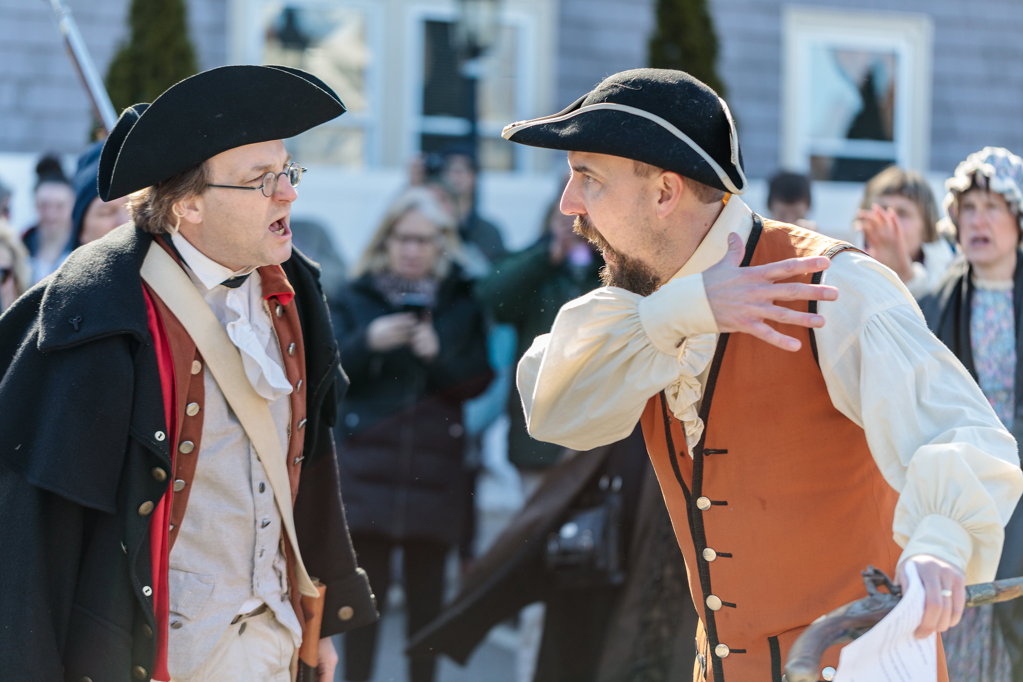Two men, dressed in historical costumes, passionately debate during a lively reenactment event, capturing the attention of an intrigued audience.