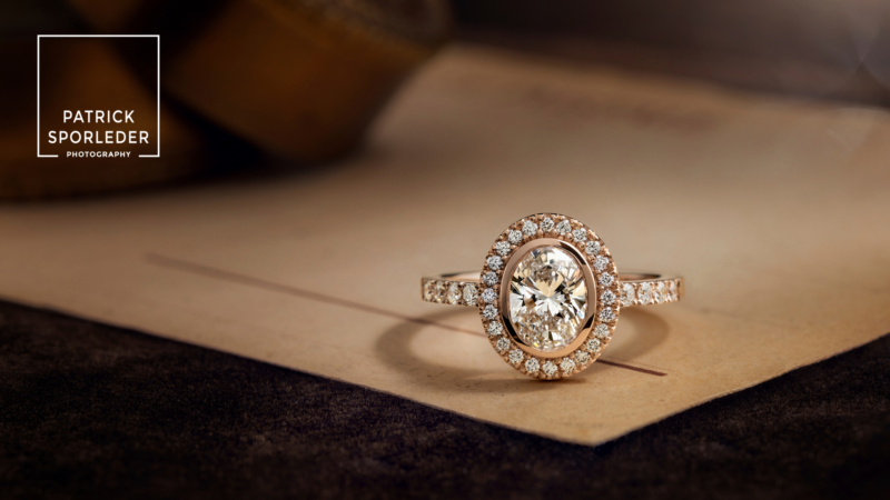 Discover the allure of an oval diamond ring, beautifully surrounded by a halo of sparkling smaller diamonds. Captured in stunning detail by Patrick Sporleder Photography, this exquisite piece rests elegantly on a delicate paper backdrop. Perfect for showcasing elegance and grace in your jewelry collection.