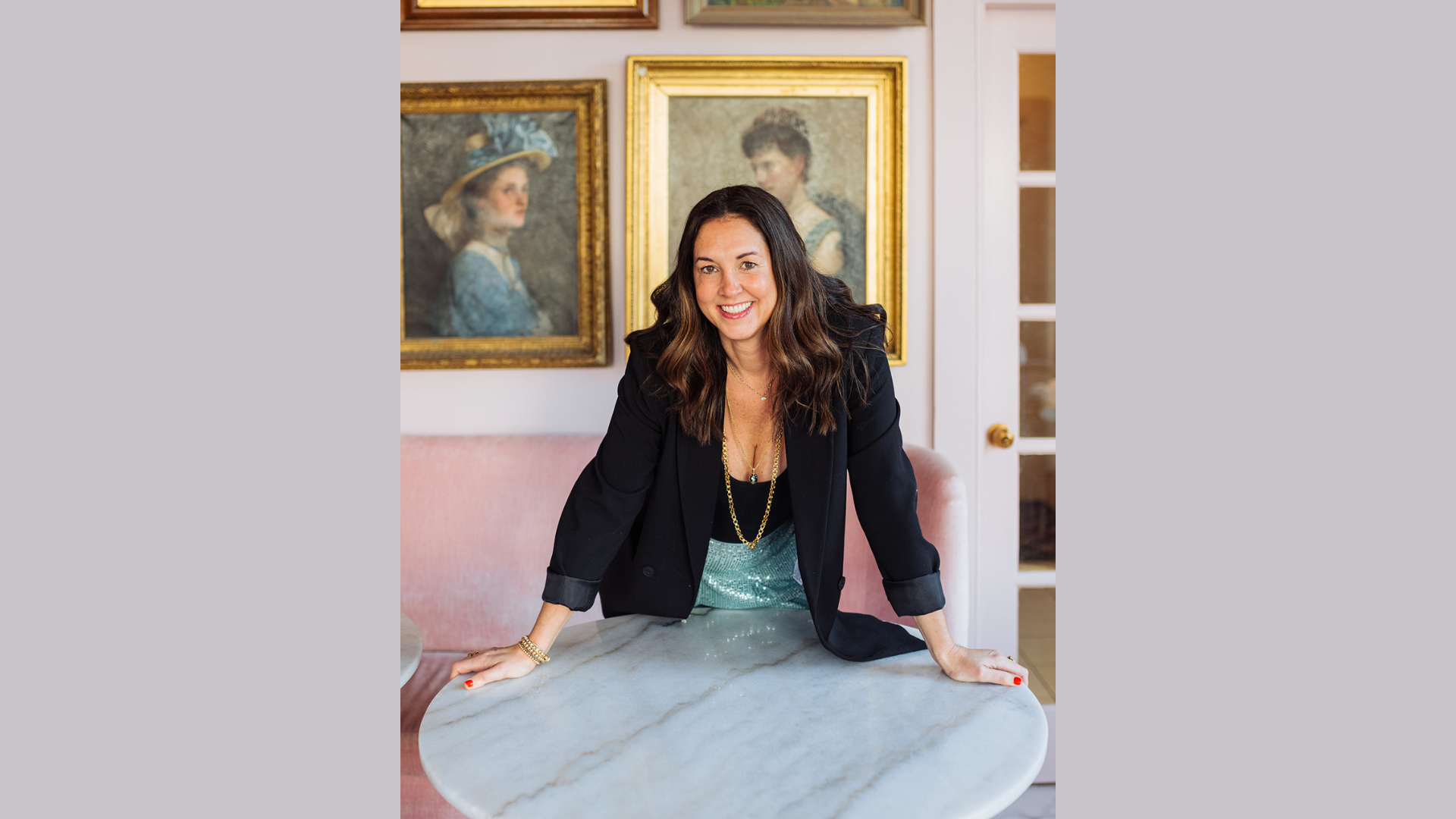 Introducing the Member: A woman in a sleek black blazer from the vibrant Wicked Ducks team leans over a marble table, with elegant framed portraits adorning the wall behind her.