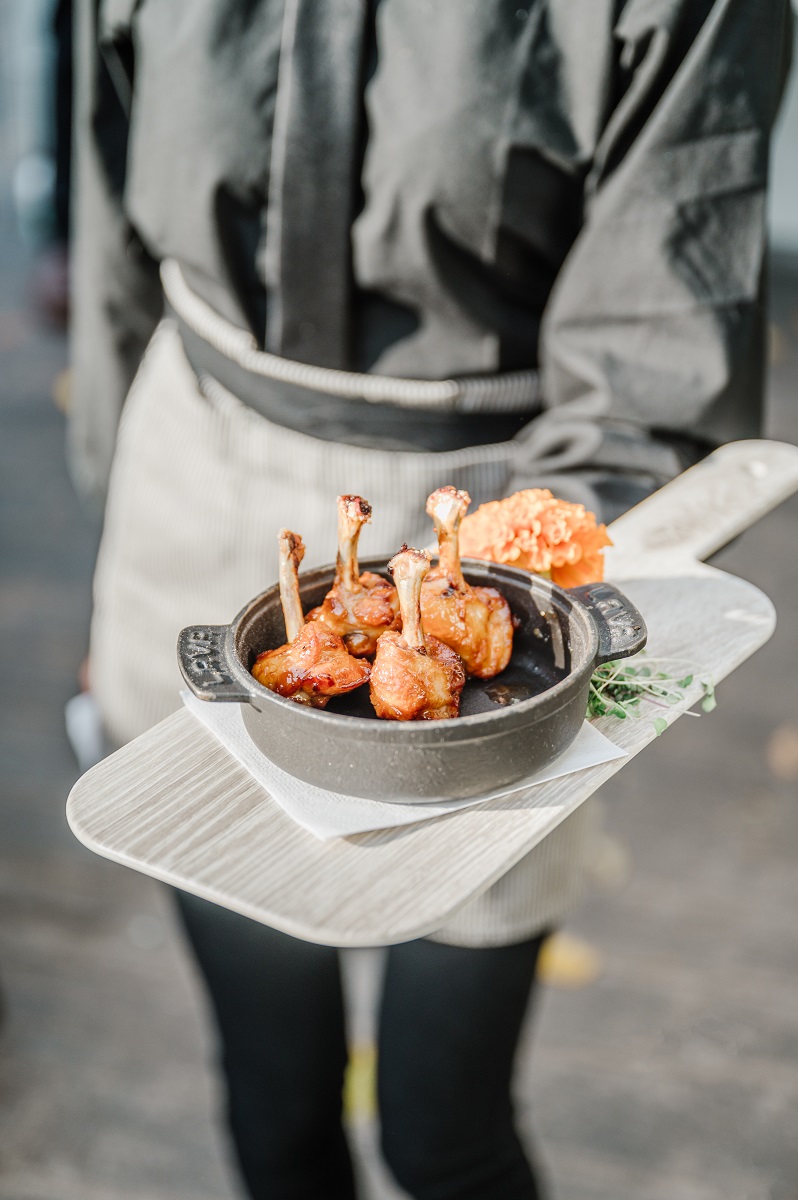 A person wearing a black shirt presents a wooden tray, showcasing a small pot filled with glazed chicken drumettes.
