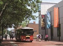 A vibrant red trolley bus glides down a historic brick street, passing a sleek modern building adorned with eye-catching banners. Nearby, people enjoy a sunny stroll under the shade of lush, leafy trees.