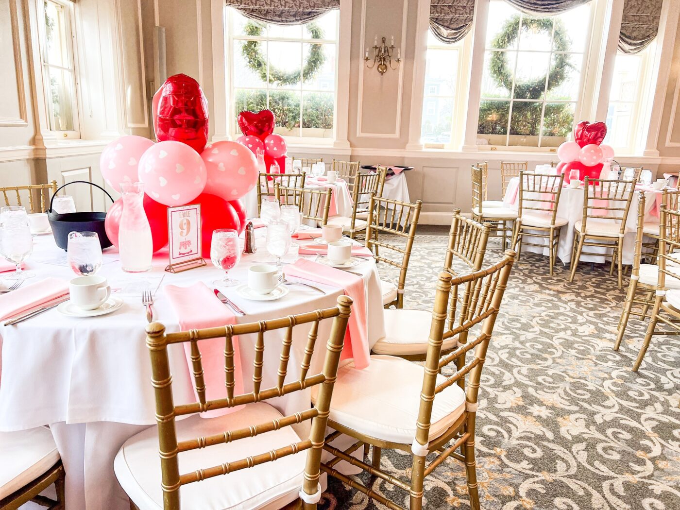 Discover our exquisite banquet hall, perfect for your next event. Picture elegant round tables adorned with vibrant pink and red balloon centerpieces, complemented by luxurious gold chairs. Floor-to-ceiling windows flood the space with natural light, creating a stunning setting for any occasion.