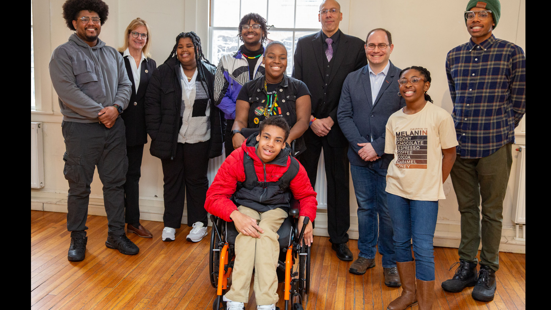 A diverse group of eight adults and one child in a wheelchair gather indoors, all smiles and dressed in a mix of casual and business attire. Their joyful presence perfectly captures the spirit of Salem's Black History Month events for 2025 as they stand on wooden flooring.