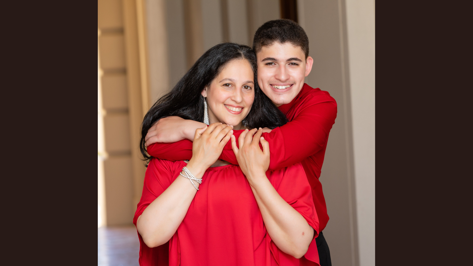 In a hallway, a woman and a young boy, both dressed in red tops, share a warm smile as the boy wraps his arms around her from behind. This genuine moment of connection perfectly mirrors the welcoming atmosphere found at "Meet the Member: Reality Lab.