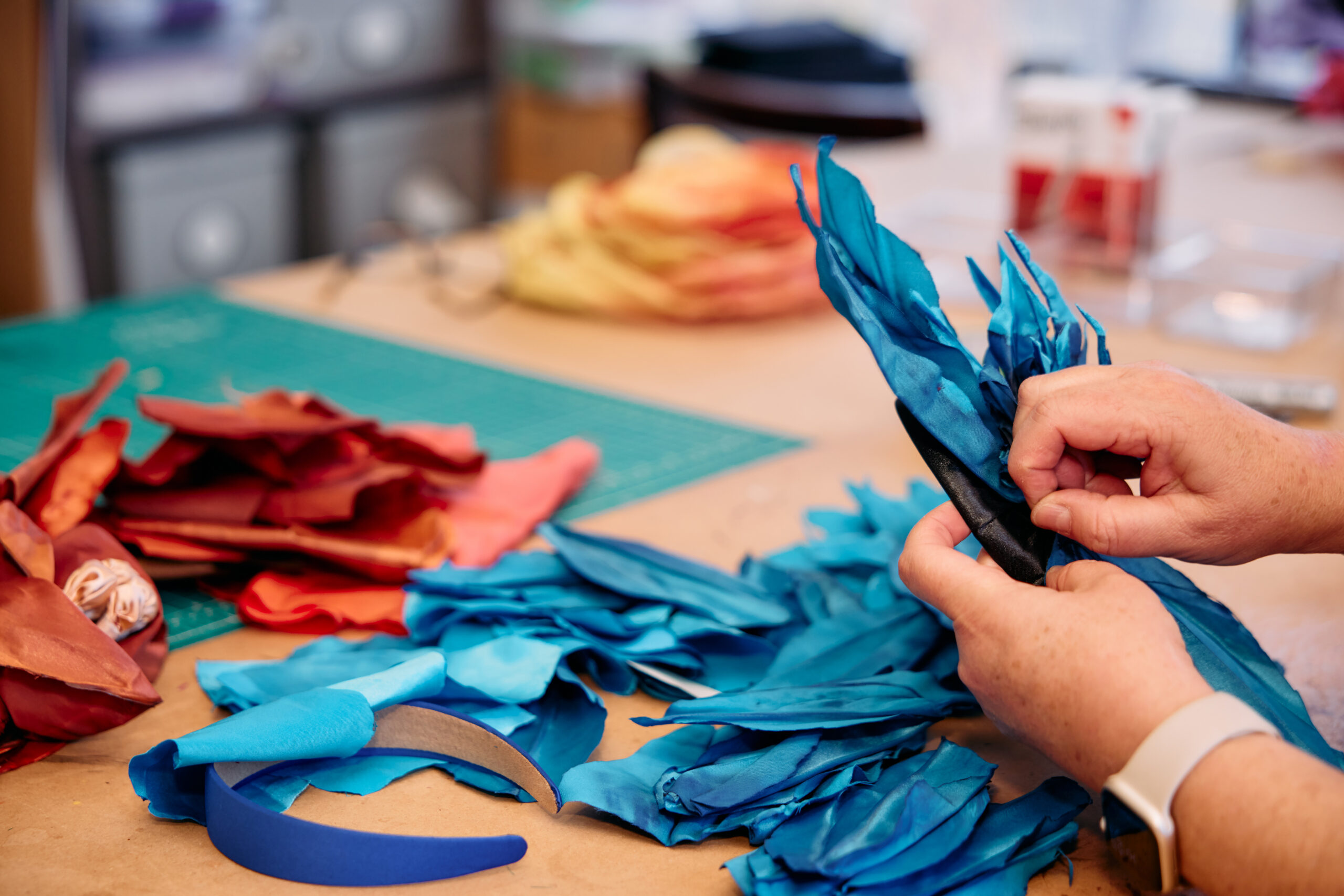 People working on blue fabric feathers at a table, surrounded by red and orange fabric feathers. In the background, there’s a cutting mat and various crafting tools.