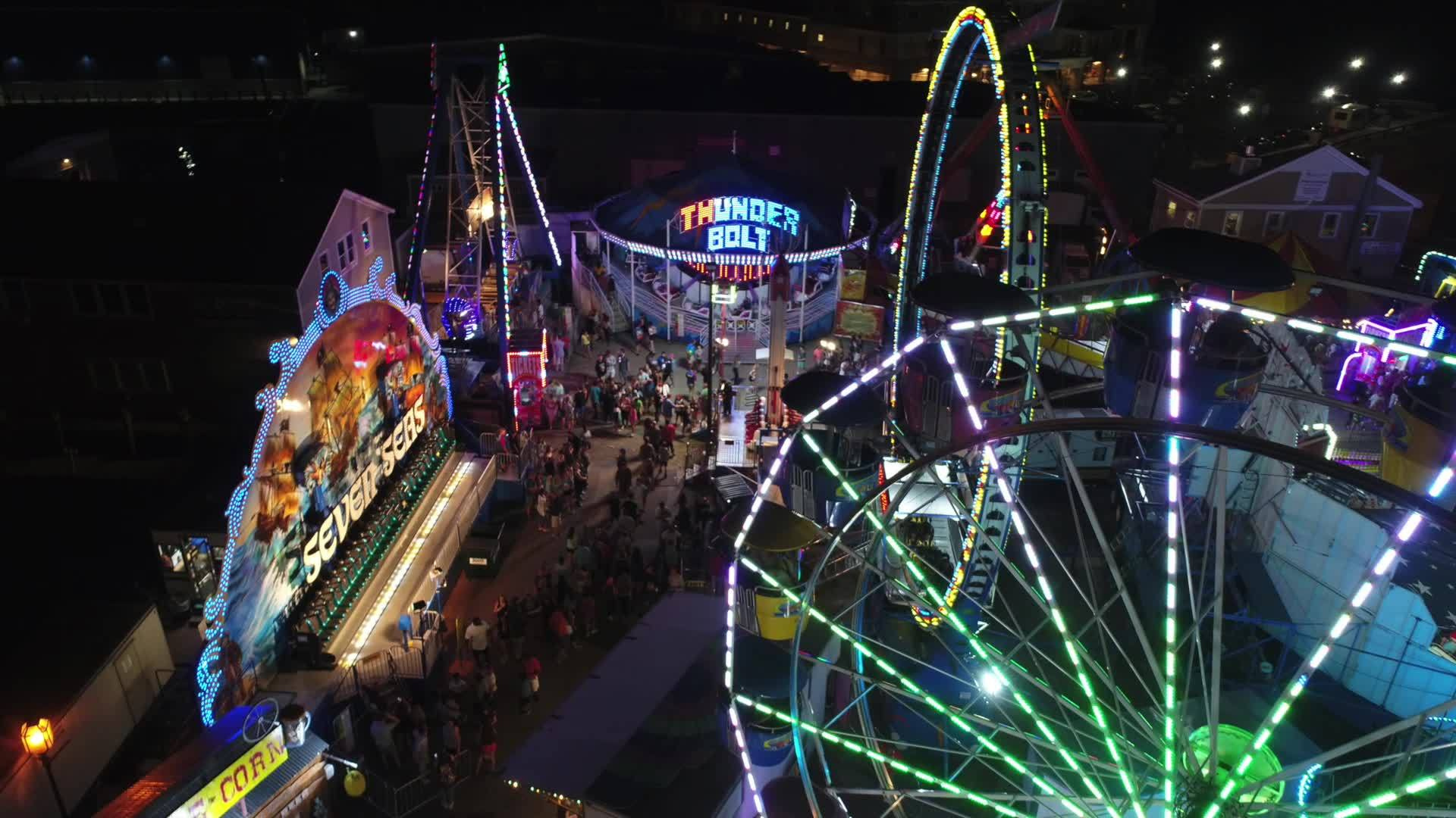 Experience the excitement of a vibrant amusement park at night from above! Picture a dazzling Ferris wheel, the thrilling Thunder Bolt ride, and the exhilarating Sea Ray, all aglow with dazzling colors. Watch crowds delight in this electrifying atmosphere—a true visual feast for thrill-seekers and night owls alike!.