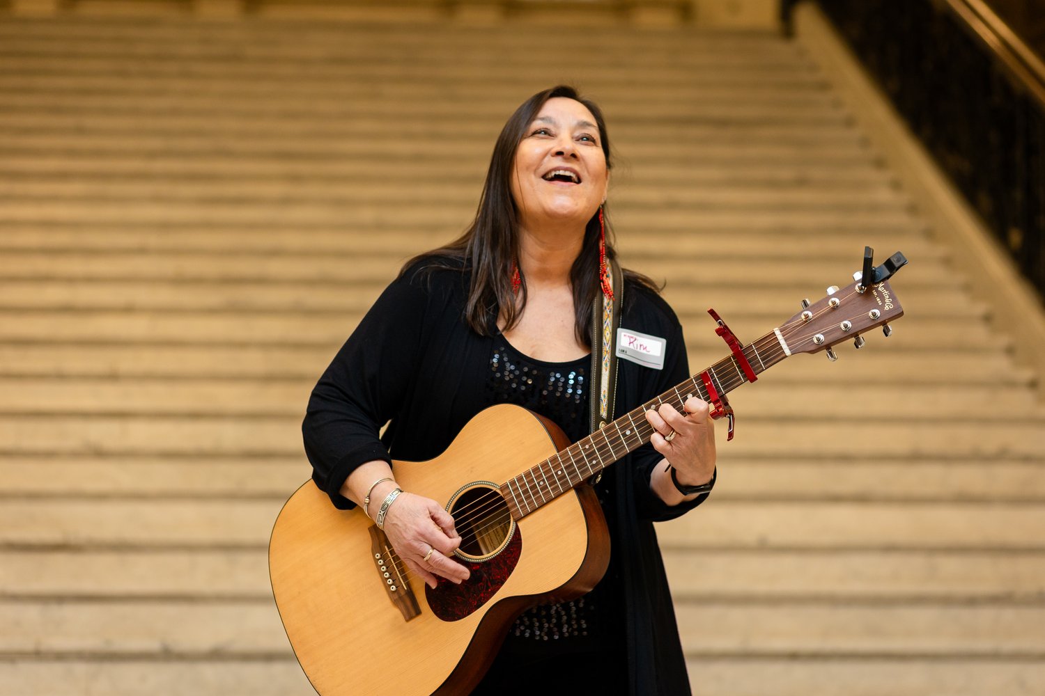 A woman in a black outfit, with a name tag, stands on the stairs playing an acoustic guitar. She's smiling and gazing upwards.
