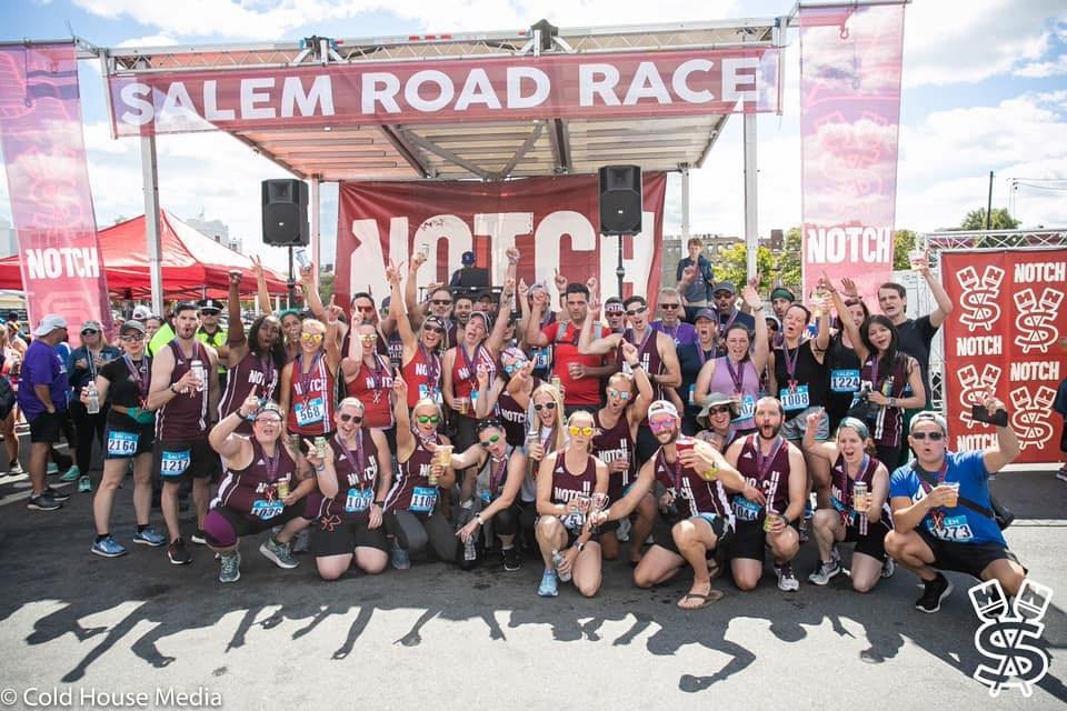 A lively group of runners celebrates together at the Salem Road Race finish line, proudly wearing maroon tank tops and race bibs while holding up their drinks. Event banners and tents can be seen in the background, capturing the fun and community spirit of this popular race.