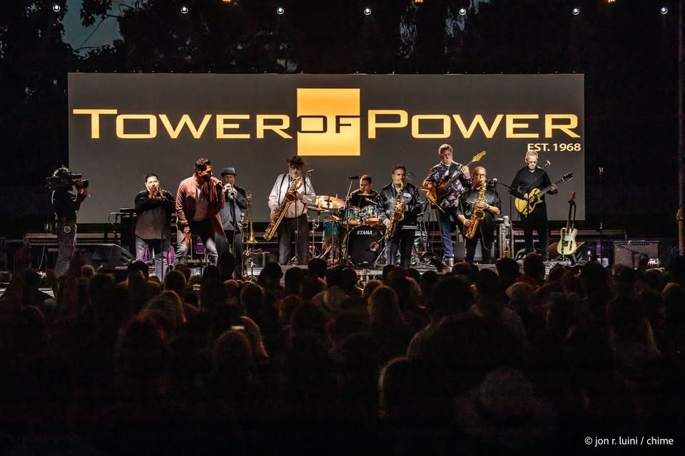 A band lights up the stage at night, captivating a crowd in front of a massive screen that reads “Tower of Power EST. 1968.” With energetic performances on brass instruments, guitar, and drums, the group delivers an unforgettable live music experience that draws fans together for a night to remember.