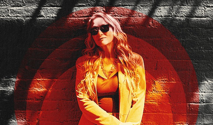 A stylish woman with long, wavy hair and sunglasses leans confidently against a brick wall decorated with bold red circular patterns. She’s wearing a light jacket and layered necklaces, creating an effortlessly chic look that captures attention.