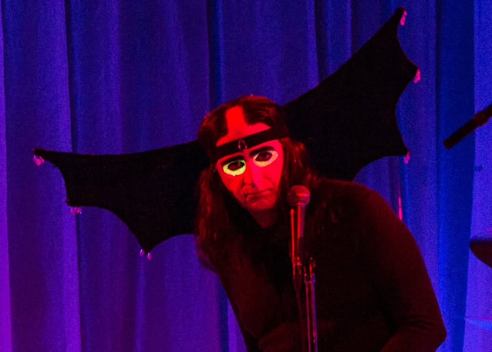 A person with long dark hair stands at a microphone, rocking glowing neon glasses and a bold headband with big bat wings. The backdrop—a vibrant purple curtain—creates an eye-catching, playful scene that’s perfect for grabbing attention in any trendy event or social media post.