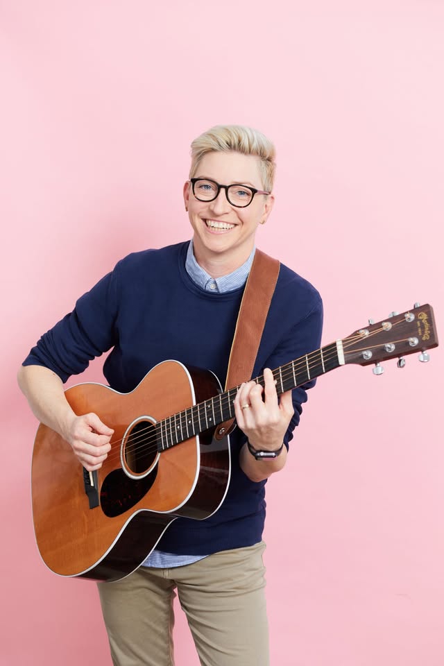 A cheerful person with short blond hair and glasses smiles as they play an acoustic guitar. Dressed in a navy sweater layered over a collared shirt and khaki pants, they stand against a vibrant pink background.