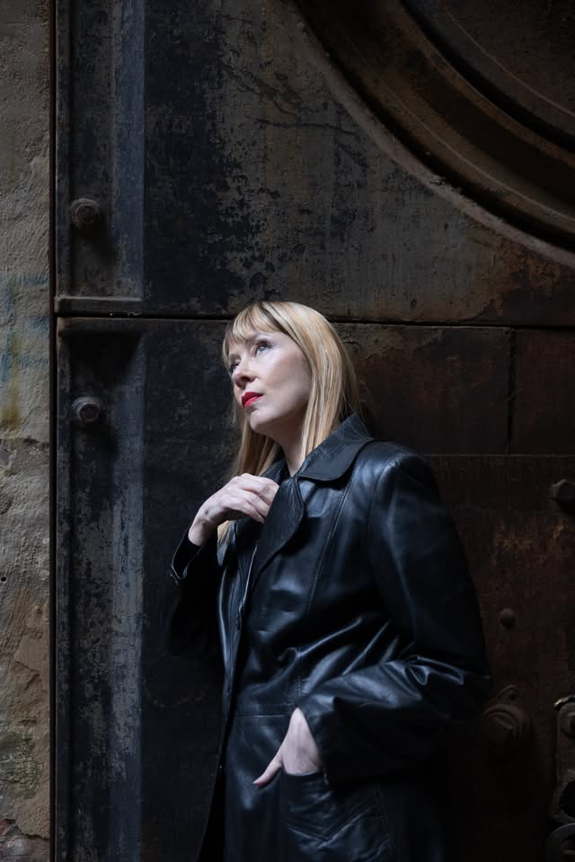 A stylish blonde woman with striking red lipstick stands in front of a weathered metal door, wearing a sleek black leather coat. She gazes upward, creating an eye-catching and thoughtful moment that brings bold fashion and urban vibes together perfectly.