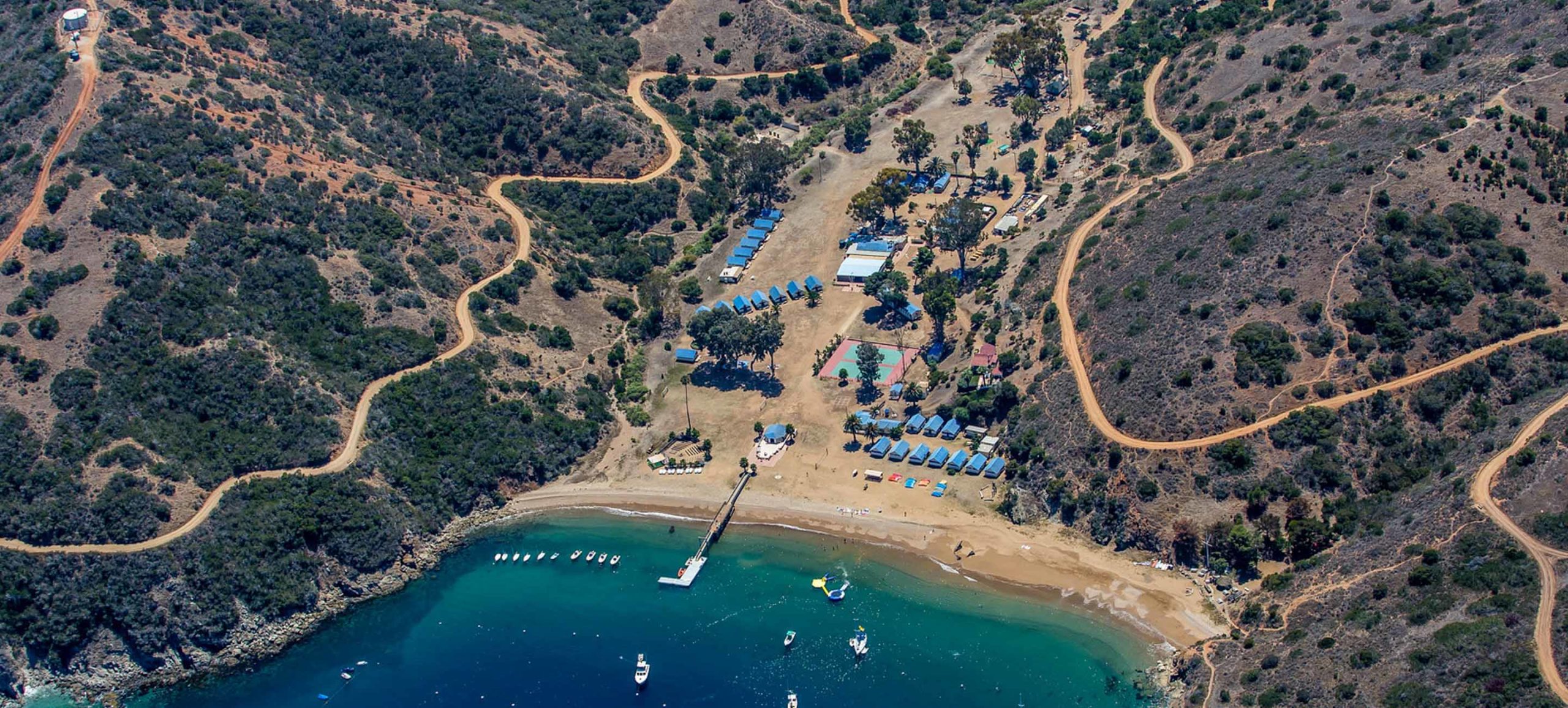 Experience the ultimate coastal getaway at our scenic camp, featuring tents and cozy cabins tucked between rolling hills. Enjoy stunning aerial views of winding dirt roads leading to a sun-kissed sandy beach, a private dock, and boats ready for adventure along the crystal-clear blue shoreline. Perfect for nature lovers seeking relaxation and outdoor fun by the sea.