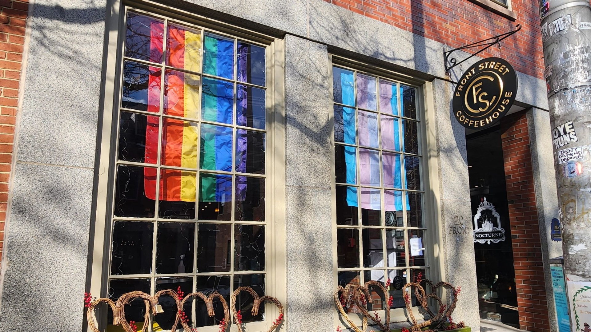 Two large windows featuring rainbow and transgender flags brighten the façade of Front Street Coffeehouse, catching the eye of passersby. Above, the café's logo is prominently displayed, while wreaths and heart decorations add a welcoming touch to the exterior.