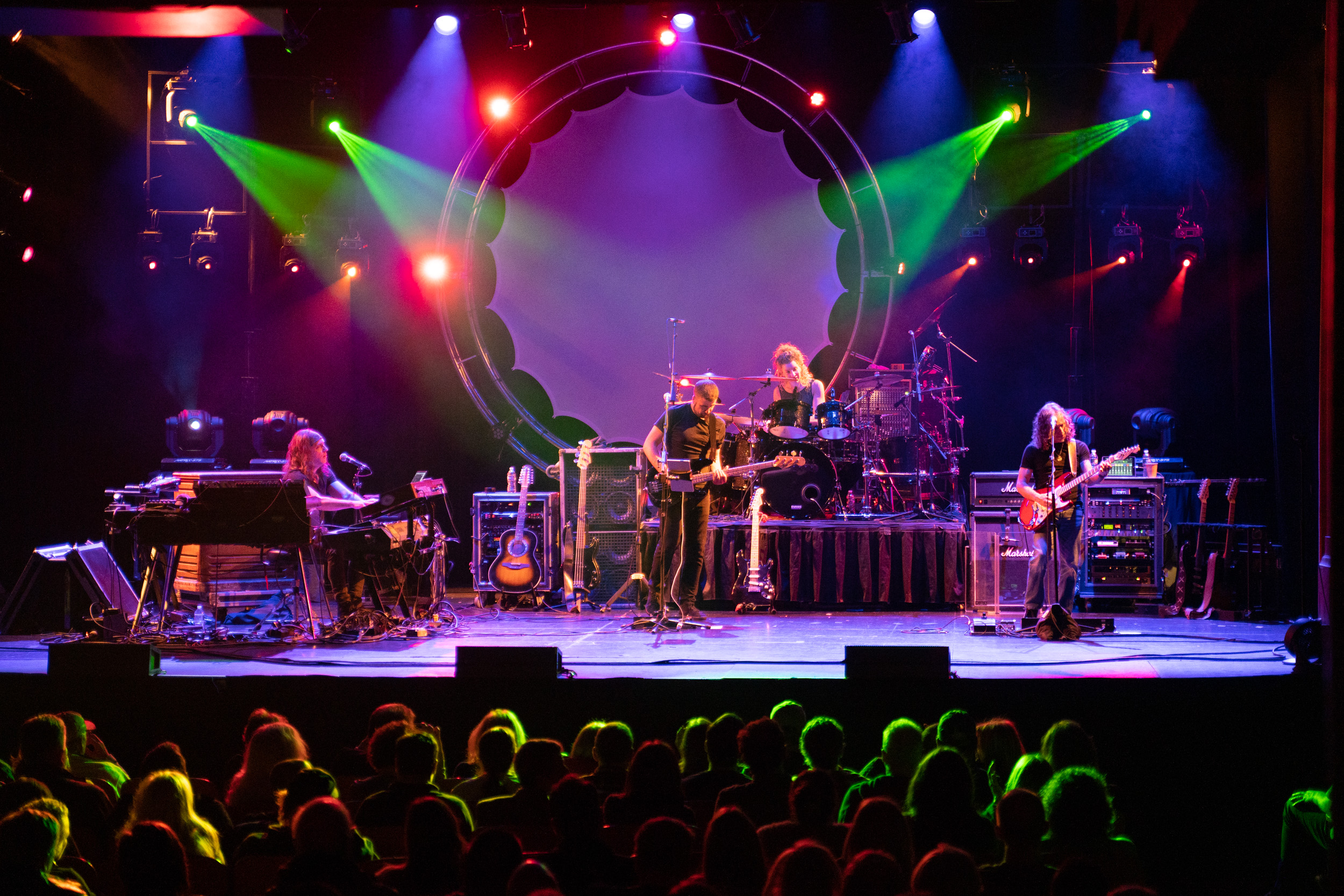 Experience the electrifying energy of a live band performance under vibrant stage lights! Watch as skilled musicians rock out with keyboards, drums, and guitars. Feel the excitement of the crowd in the foreground, sharing in this unforgettable musical journey.
