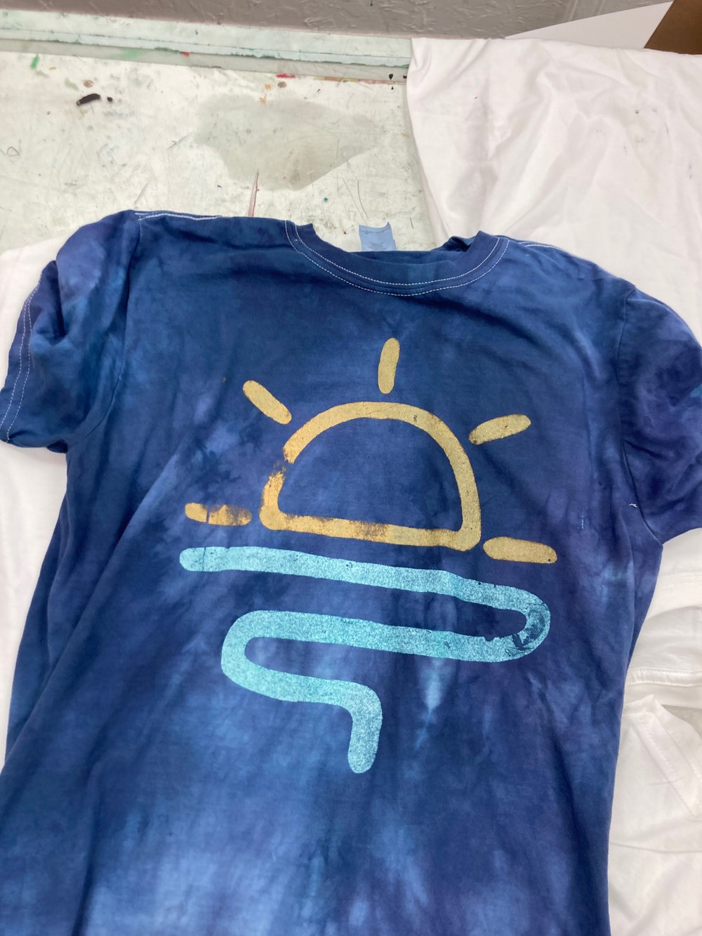 Show off your laid-back style with this blue tie-dye T-shirt, featuring a bold yellow sun and a playful blue wavy line design across the front. Perfect for sunny days, this eye-catching tee looks great laid out on a crisp white background.
